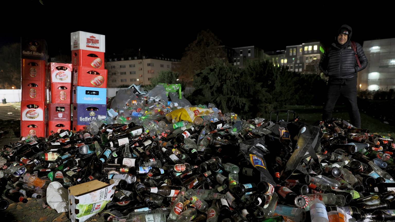 Ein Berg aus Glasflaschen liegt auf der Wiese, dahinter drei Türme aus Bierkästen. Rechts daneben steht eine Person. Im Hintergrund sieht man ein Gebäude der Universität zu Köln.