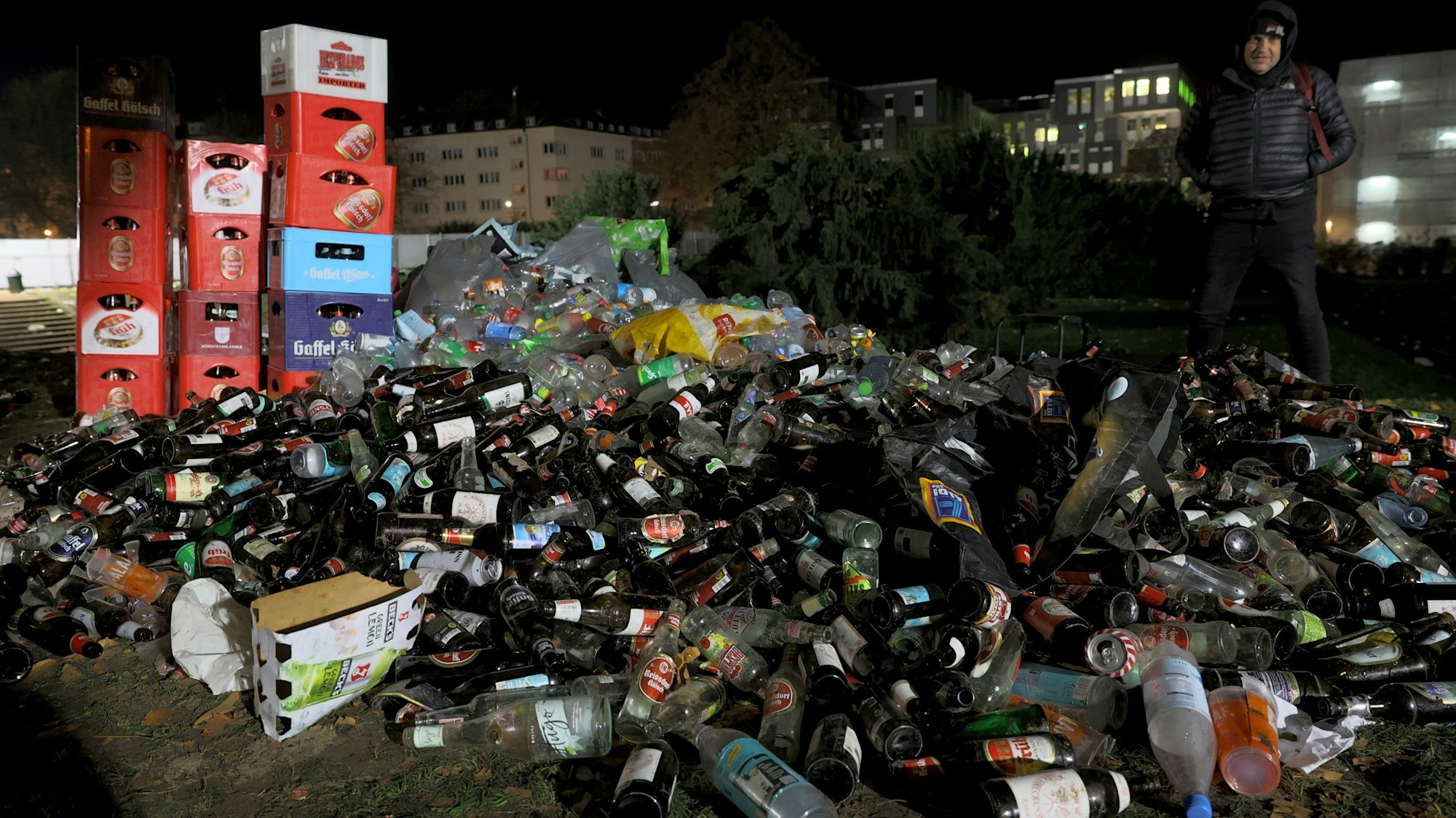 Ein Berg aus Glasflaschen liegt auf der Wiese, dahinter drei Türme aus Bierkästen. Rechts daneben steht eine Person. Im Hintergrund sieht man ein Gebäude der Universität zu Köln.
