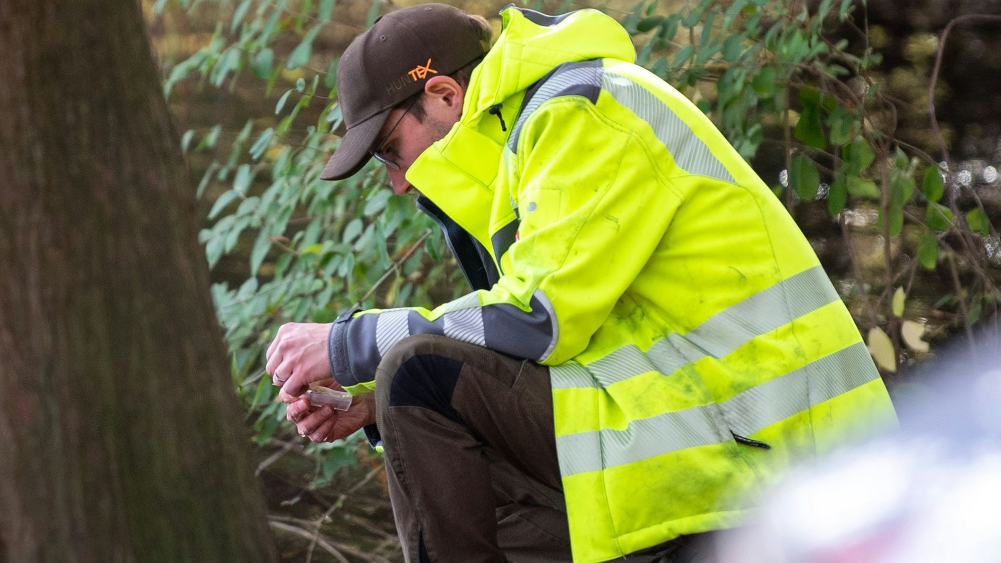Ein Mitarbeiter der Unteren Wasserbehörde kniet auf dem Boden und entnimmt eine Wasserprobe. Er trägt eine gelbe Jacke.