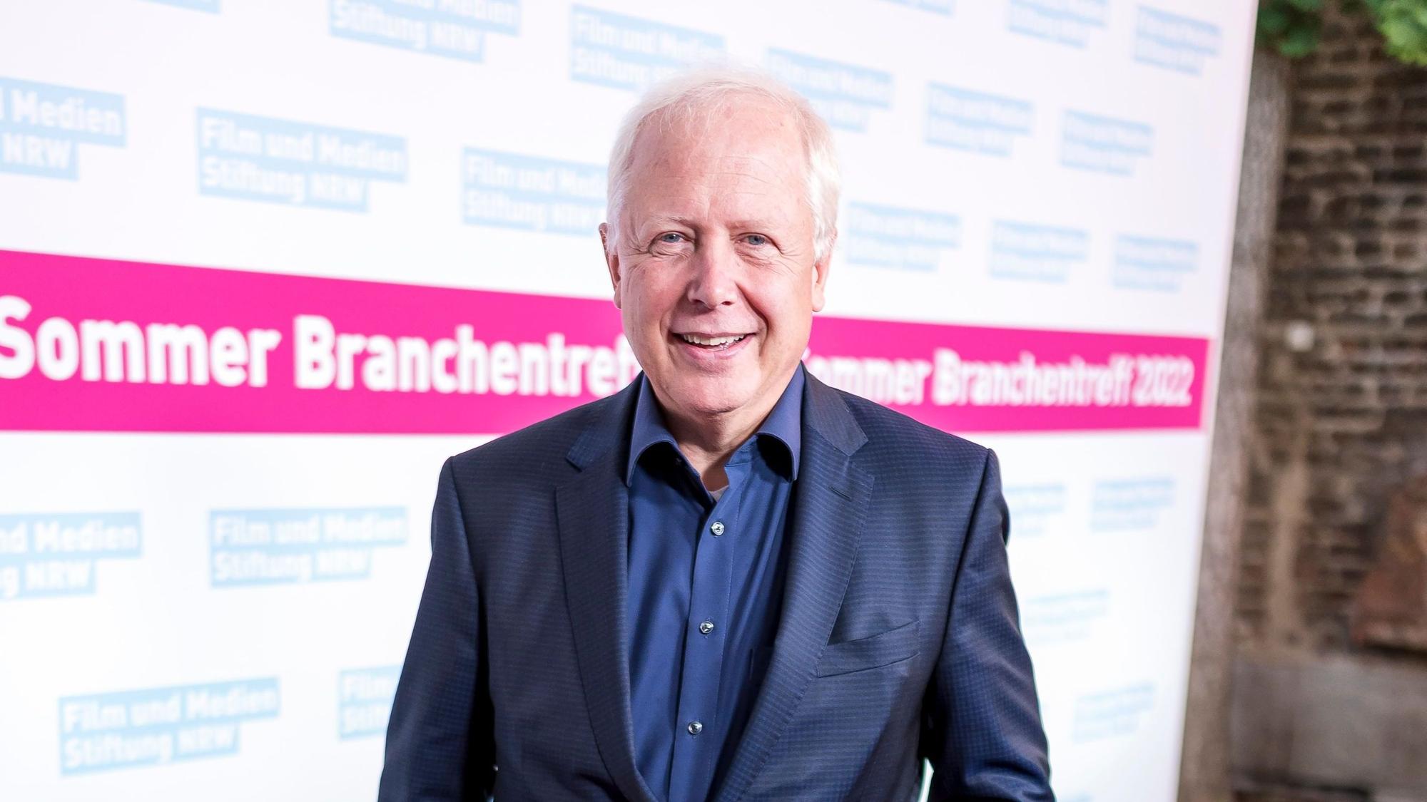 Tom Buhrow steht mit einem blauen Anzug und blauem Hemd vor einer Wand, auf der zu lesen ist „Sommer Branchentreff 2022“.