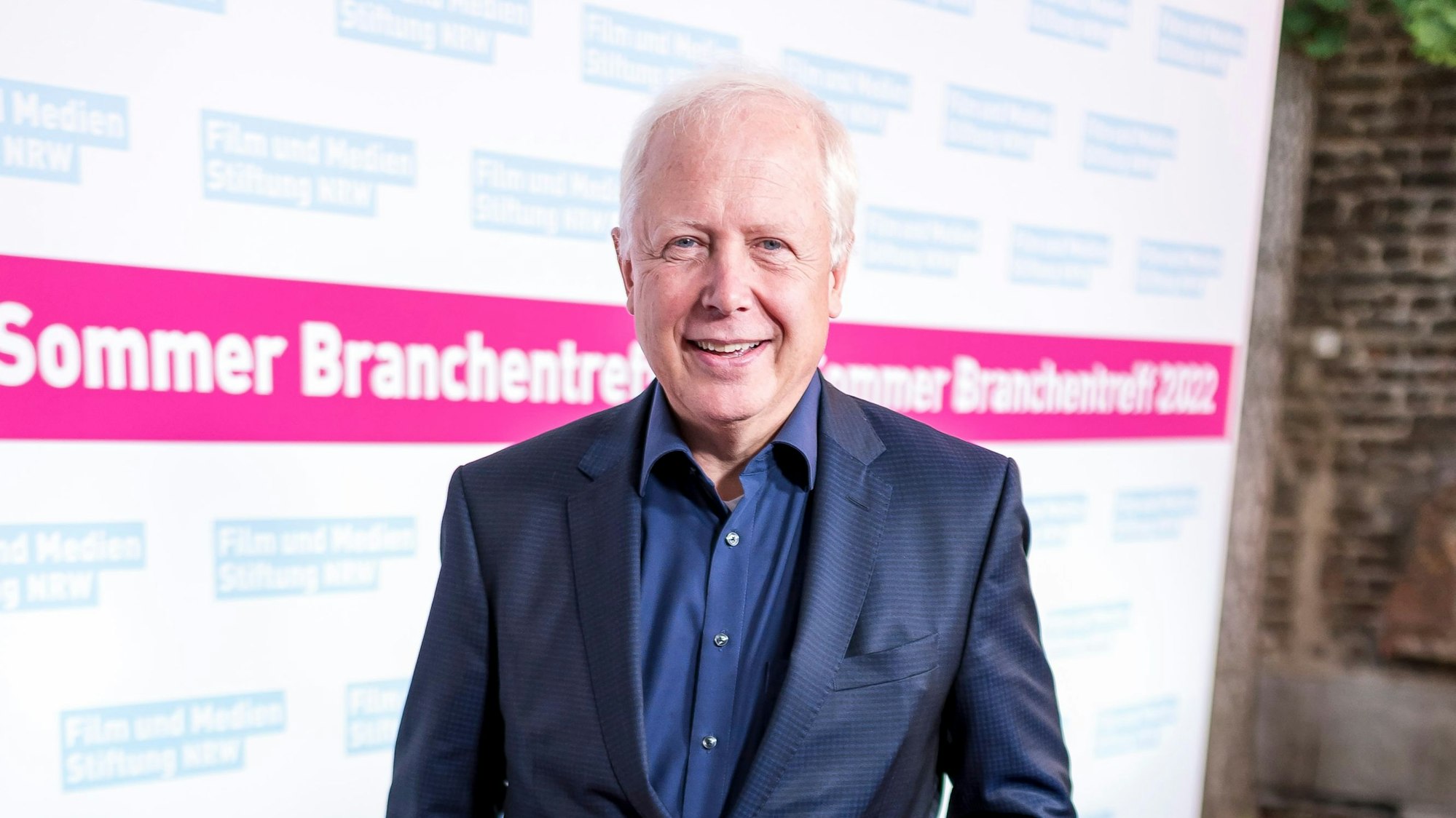 Tom Buhrow steht mit einem blauen Anzug und blauem Hemd vor einer Wand, auf der zu lesen ist „Sommer Branchentreff 2022“.