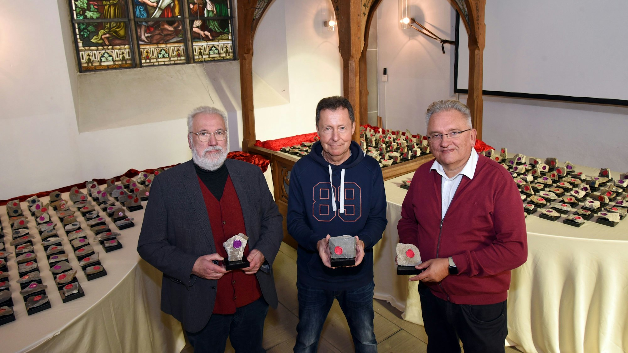 Die Pfarrer Uwe Selbach (l.) und Markus Aust (r.) sowie Rüdiger Hockamp halten drei der Unikate in den Händen. Im Hintergrund sind die weiteren Unikate auf Tischen zu sehen.