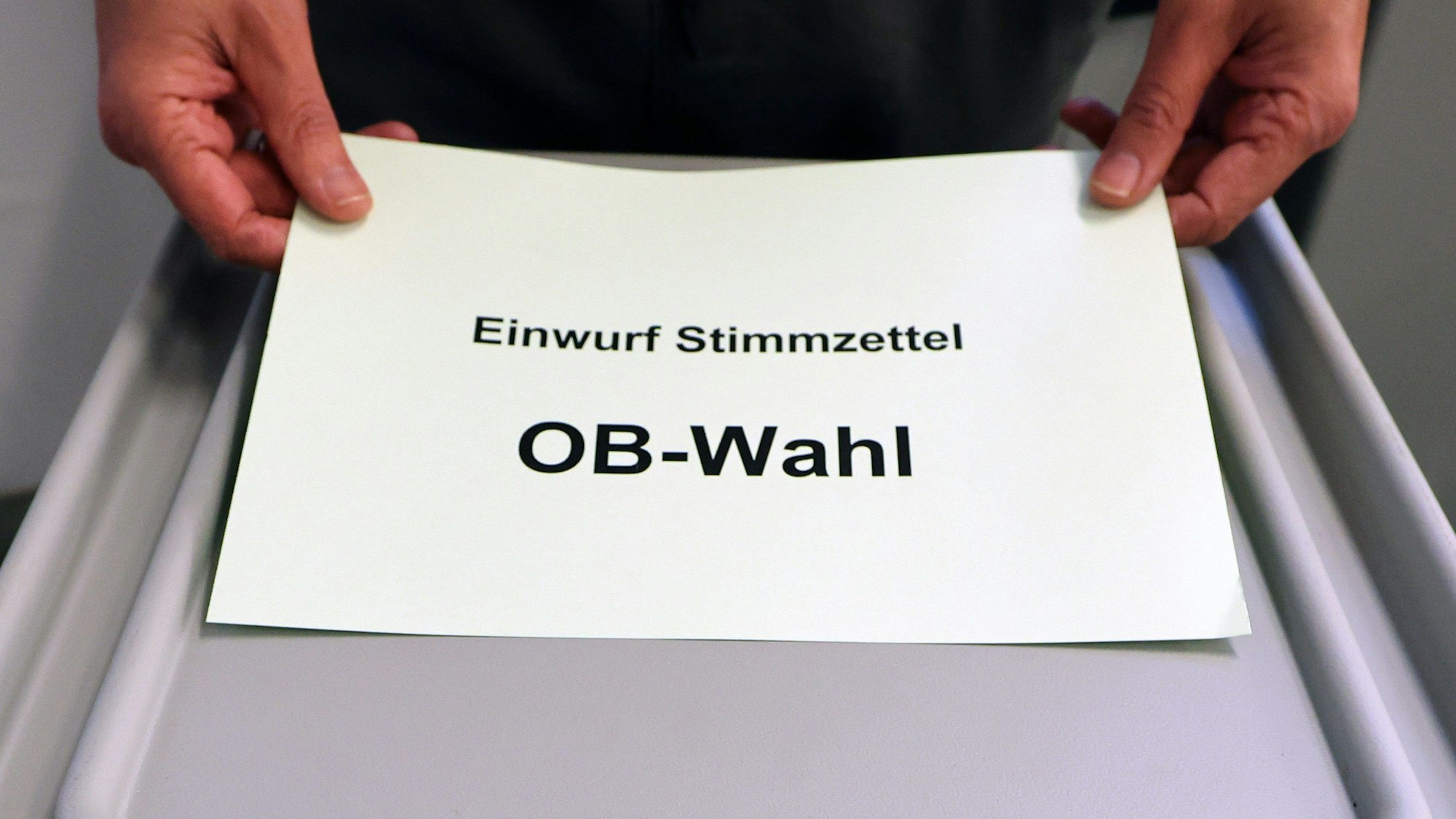 Ein Zettel mit der Aufschrift „Einfwurf Stimmzettel OB-Wahl“ wird vor einer grauen Wahlurne gezeigt.