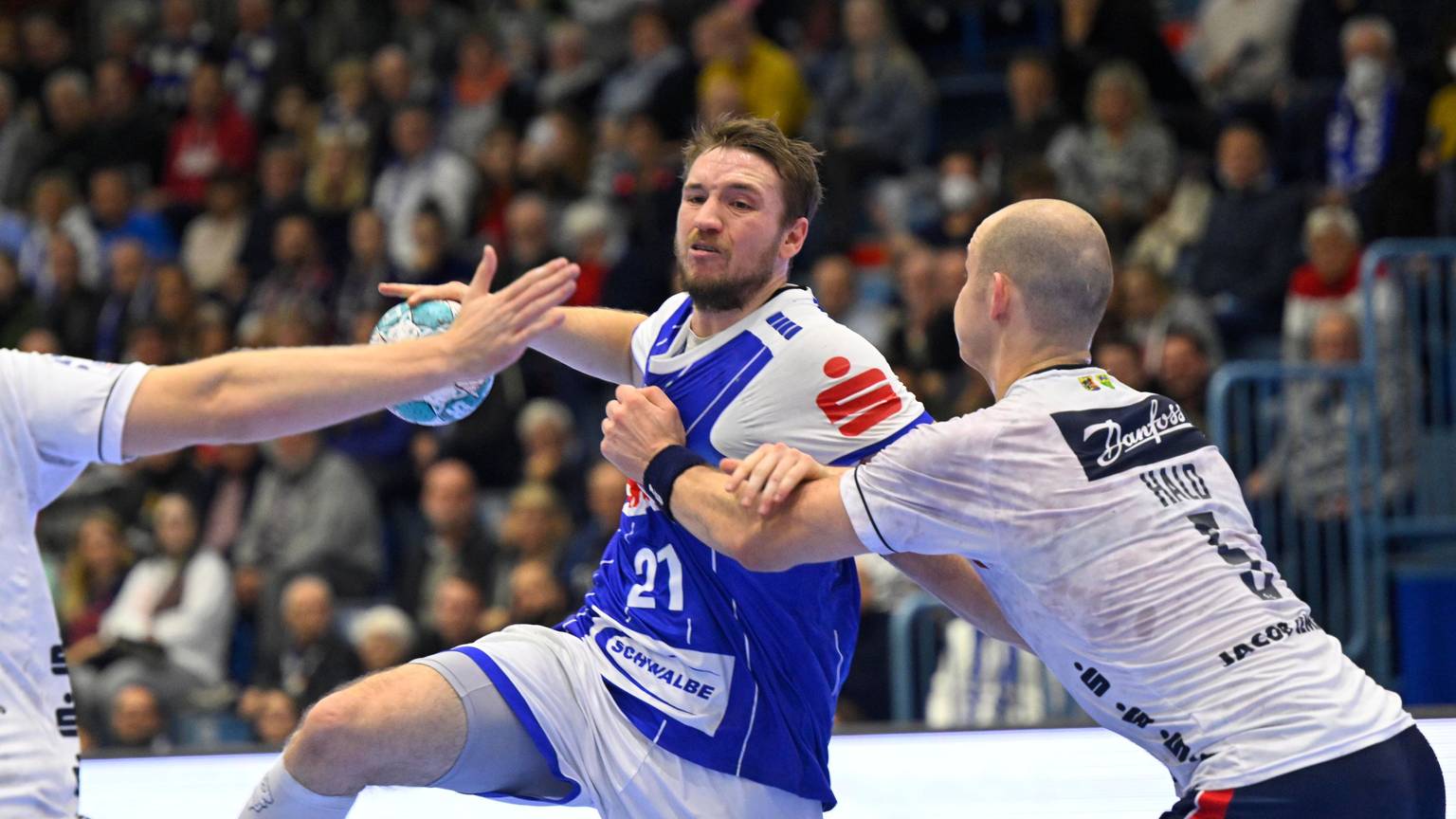 Dominik Mappes vom VfL Gummersbach trägt ein blaues Trikot. Er hält den Ball in der Hand und wird von zwei Spielern der SG Flensburg-Handewitt verteidigt, die weiße Trikots tragen.