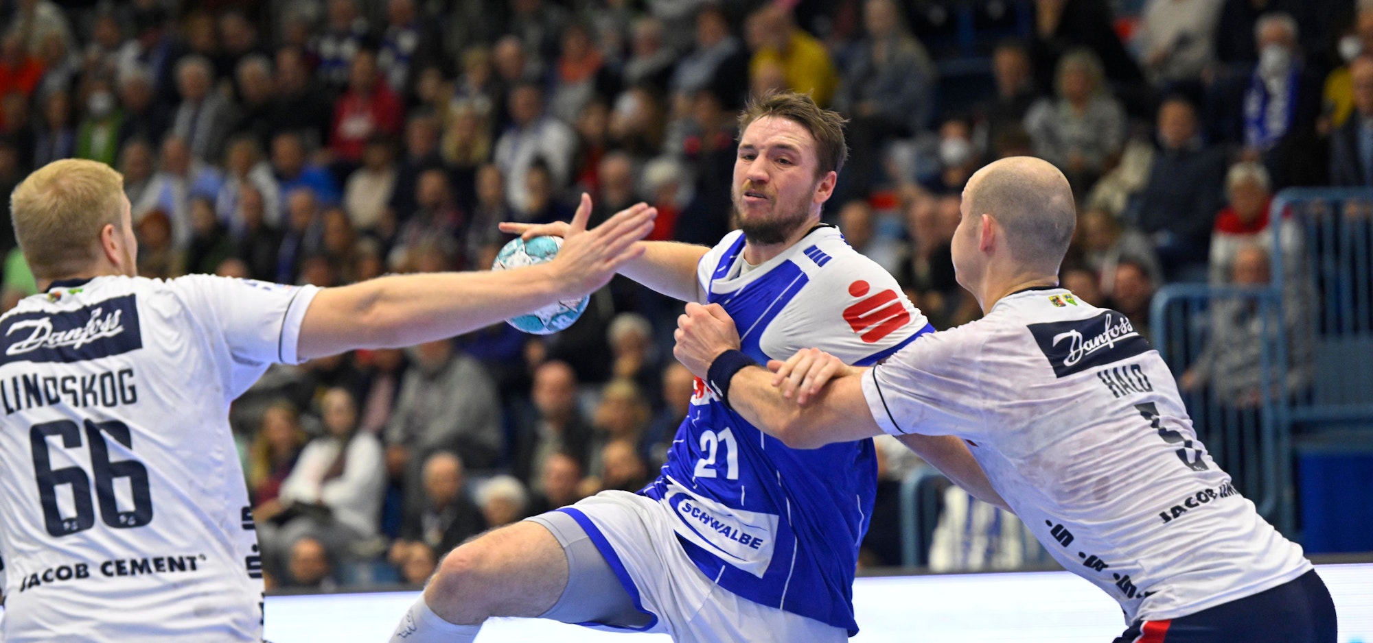 Dominik Mappes vom VfL Gummersbach trägt ein blaues Trikot. Er hält den Ball in der Hand und wird von zwei Spielern der SG Flensburg-Handewitt verteidigt, die weiße Trikots tragen.
