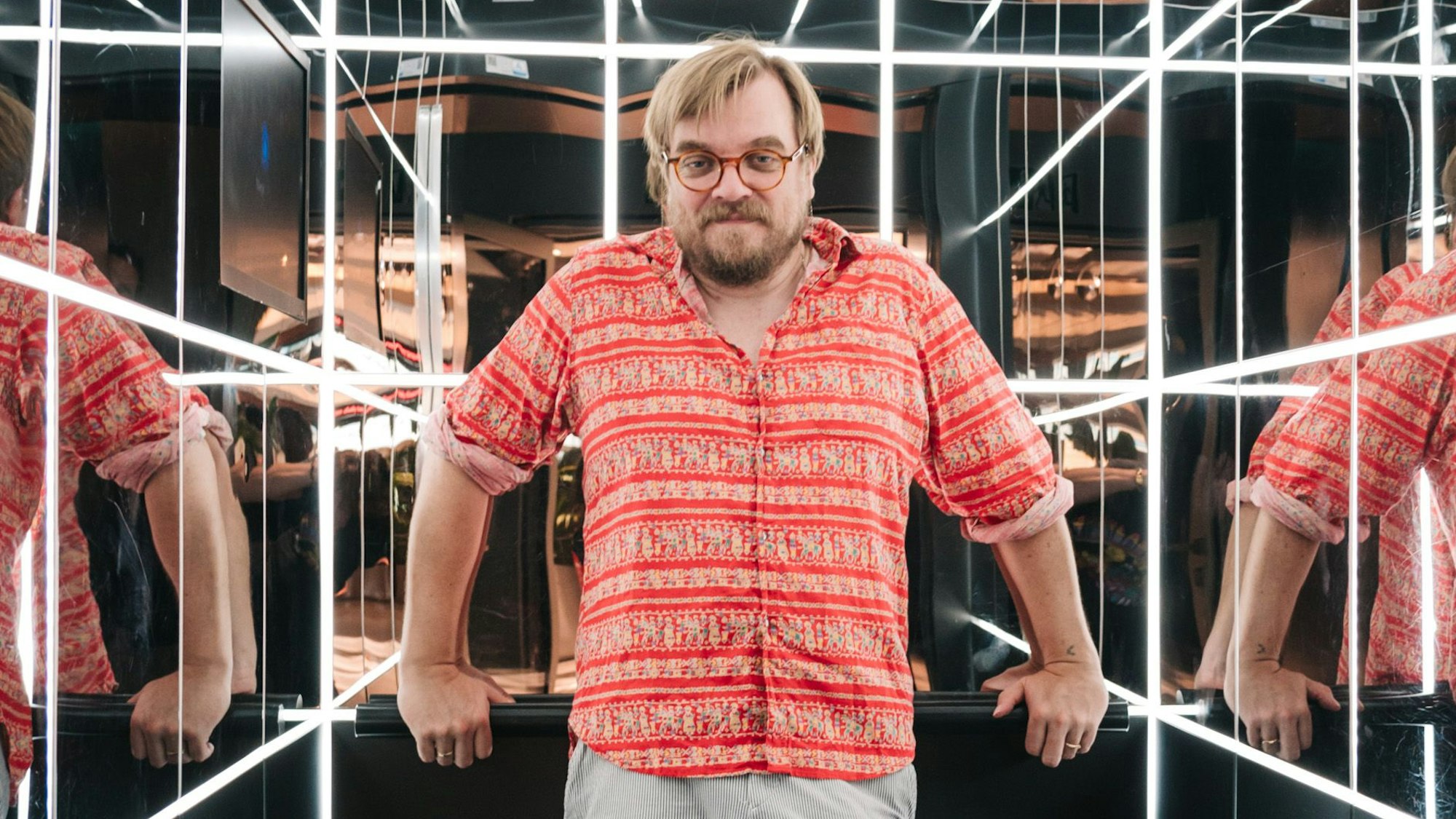 Nilz Bokelberg steht in einem verspiegelten Aufzug mit Leuchtröhren.