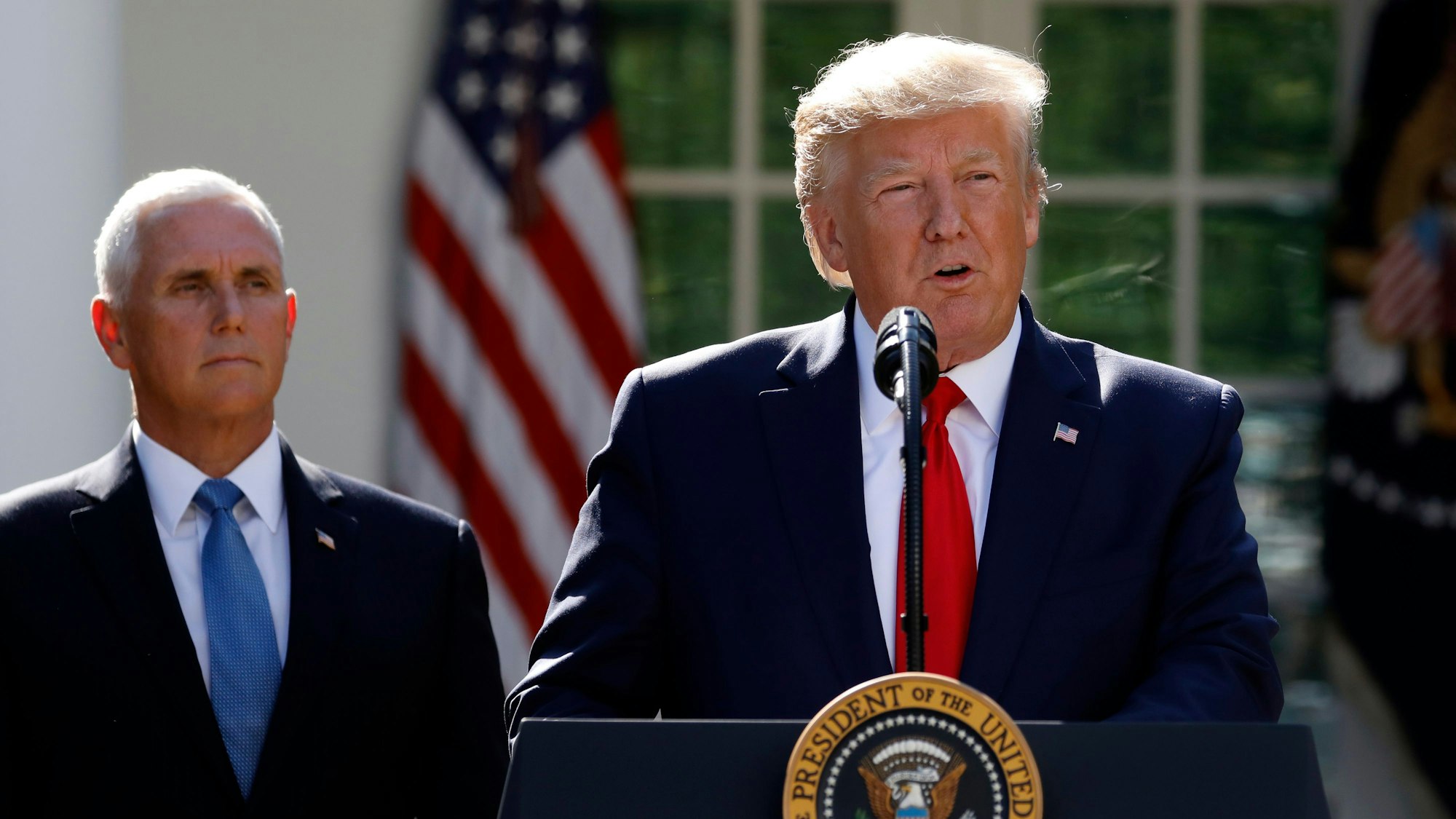 Der ehemalige US-Präsident Donald Trump steht während seiner Amtszeit an einem Mikrofon und spricht. Hinter ihm steht Mike Pence.