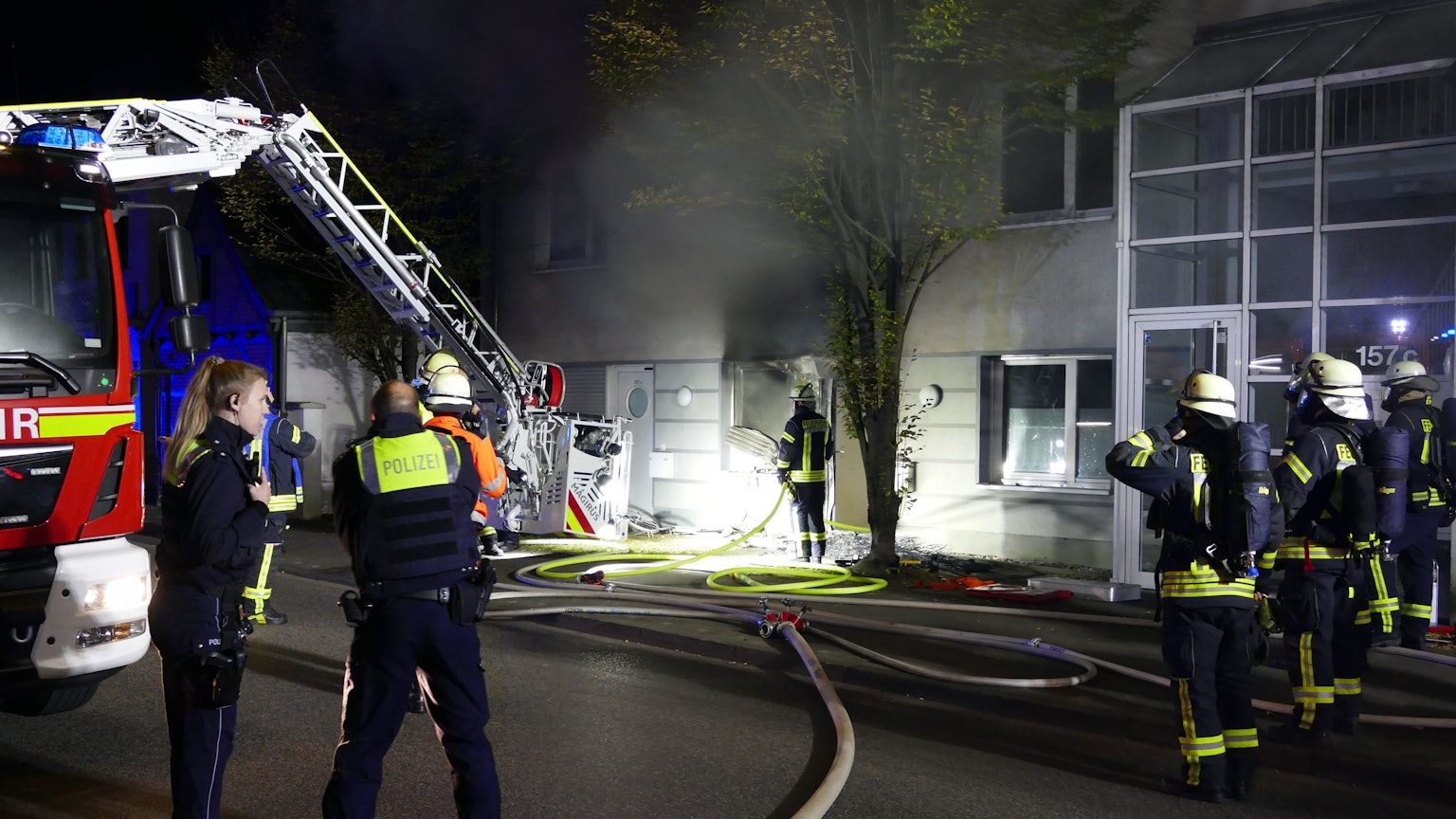 Feuerwehrleute stehen in der Nacht vor einem Fenster, aus dem dicker, schwarzer Rauch quillt. Ein Feuerwehrwagen steht daneben, ein Schlauch liegt auf der Straße.