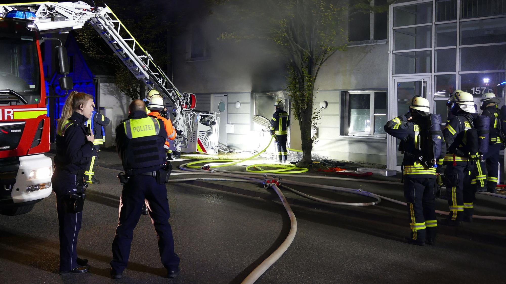 Feuerwehrleute stehen in der Nacht vor einem Fenster, aus dem dicker, schwarzer Rauch quillt. Ein Feuerwehrwagen steht daneben, ein Schlauch liegt auf der Straße.