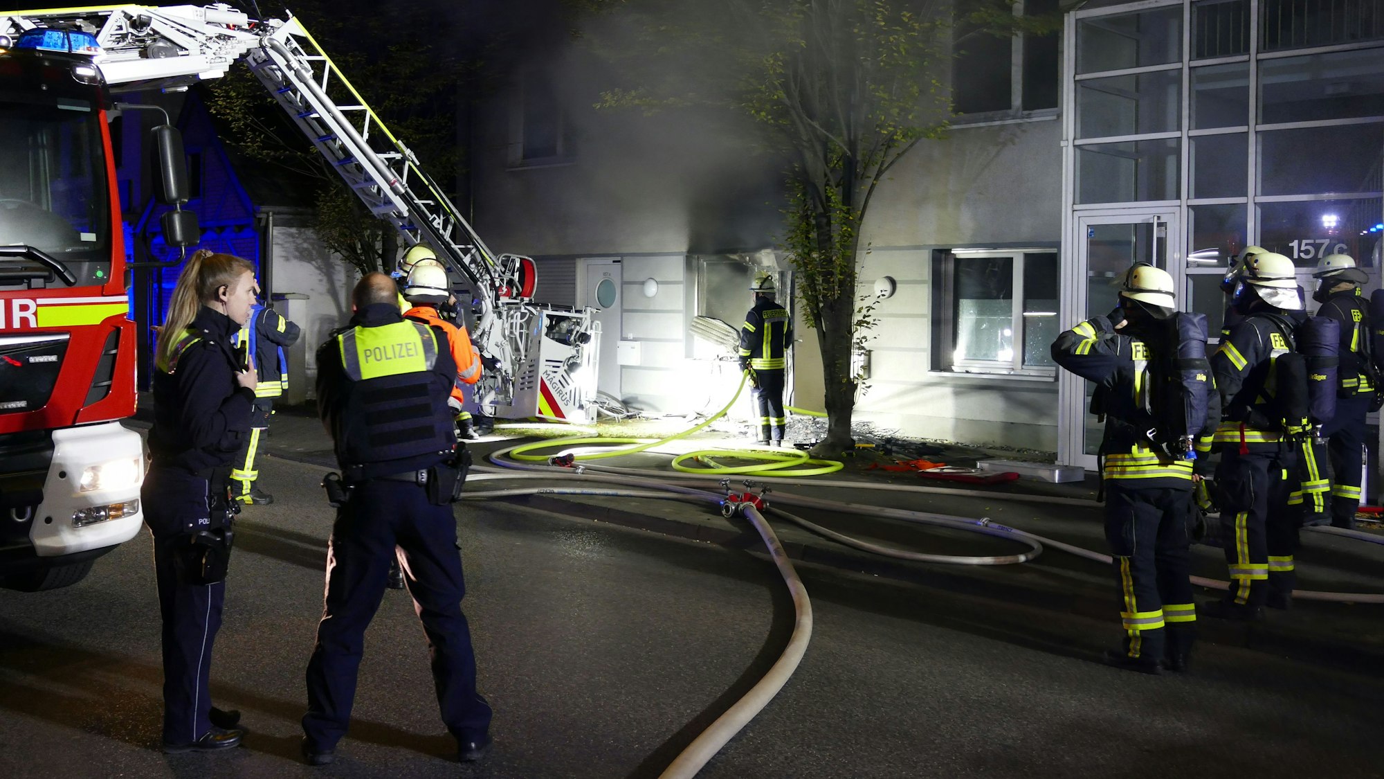Feuerwehrleute stehen in der Nacht vor einem Fenster, aus dem dicker, schwarzer Rauch quillt. Ein Feuerwehrwagen steht daneben, ein Schlauch liegt auf der Straße.