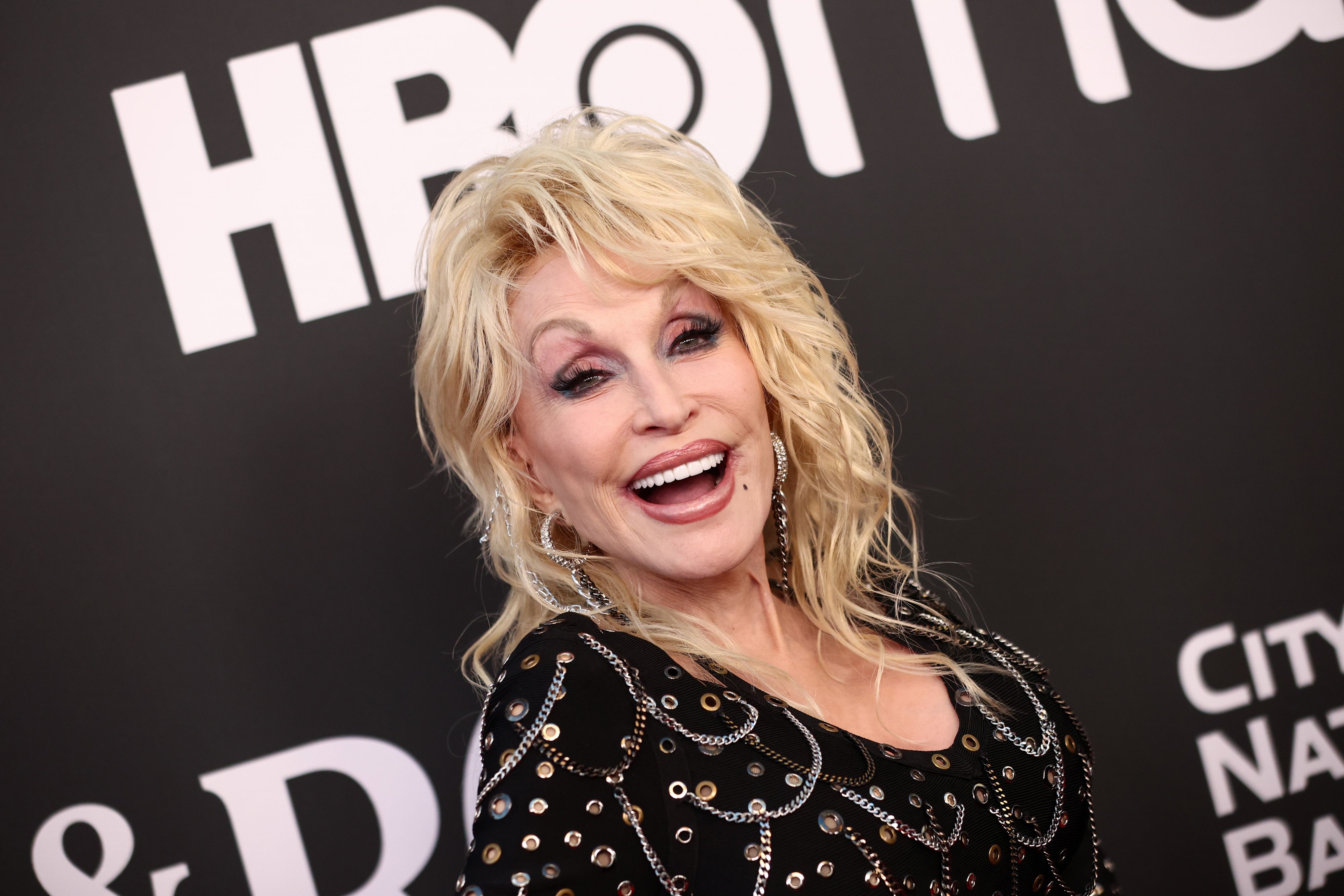 Dolly Parton lacht vor einem HBO-Schild für die Fotografen auf dem roten Teppich.
