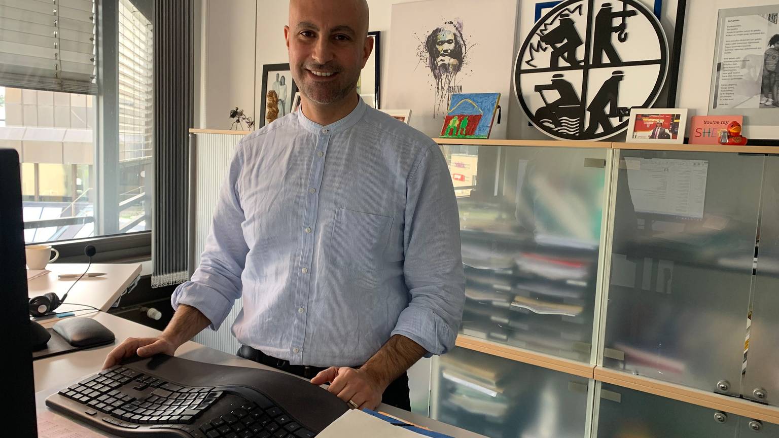 Der SPD-Politiker Ali Dogan trägt ein blaues Hemd und steht in seinem Büro im Rathaus von Sankt Augustin.