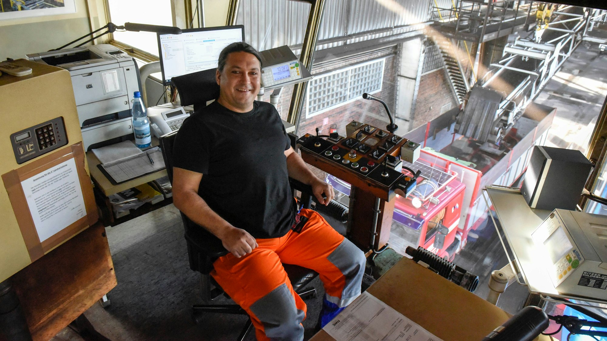 Ein Mann sitzt auf einem Stuhl und hat vor sich mehrere Knöpfe. Durch eine Scheibe kann er auf die LKW blicken, die mit Kohle beladen werden.