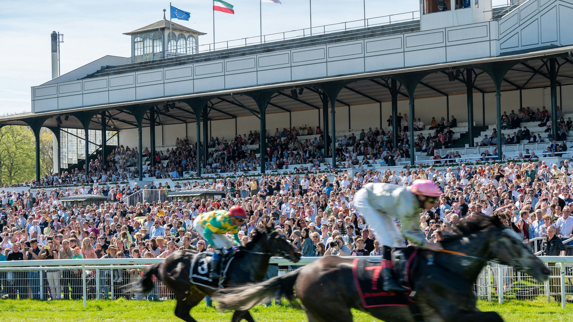 Zwei Reiter preschen an der Tribüne der Pferderennbahn Weidenpesch vorbei.
