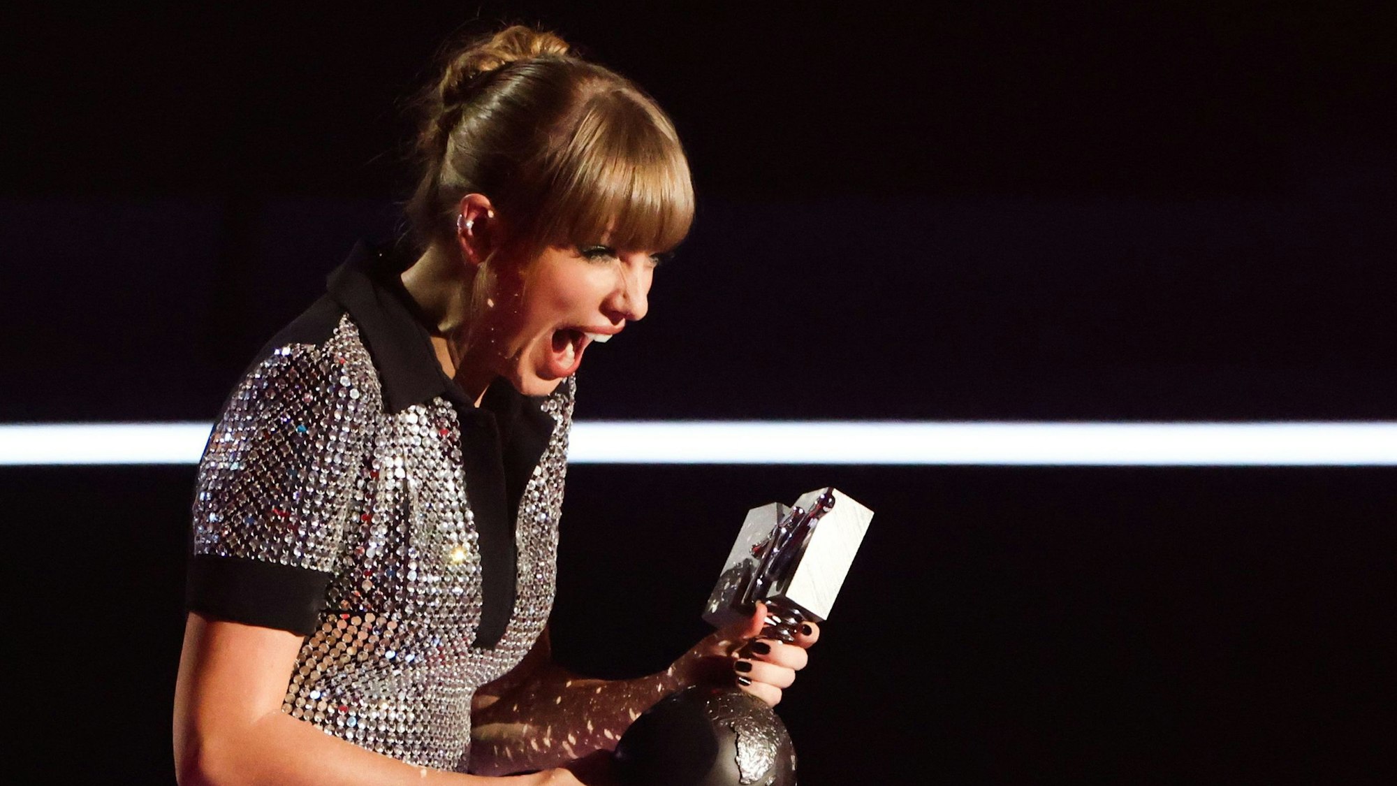 Taylor Swift und ihre Fans freuen sich über vier Preise. Die Sängeri reist vor Freunde den Mund auf und schreit. Sie trägt eine Glitzerbluse im Discokugel-Stil. Sie hält die MTV-Trophäe in der Hand.