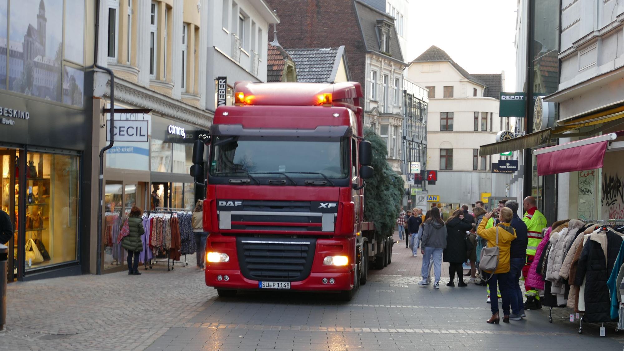 Der Lkw fährt durch die Fußgängerzone. Die Lkw liegt auf dem Anhänger. Eine Frau macht mit ihrem Handy ein Bild von der Szene.