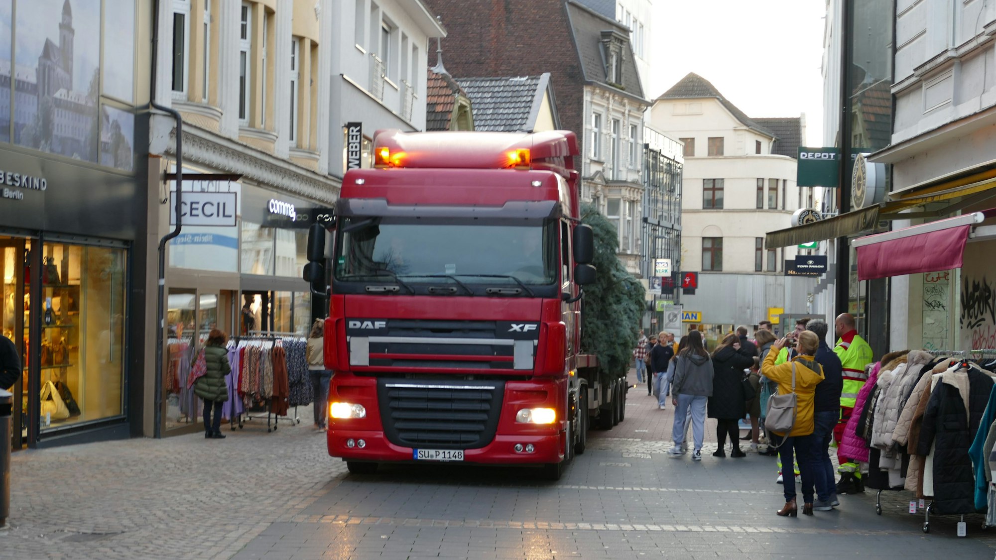 Der Lkw fährt durch die Fußgängerzone. Die Lkw liegt auf dem Anhänger. Eine Frau macht mit ihrem Handy ein Bild von der Szene.