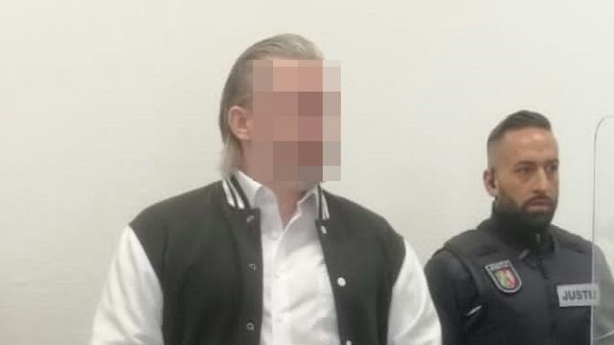 Der Angeklagte steht hinter einem Stuhl im Gerichtssaal. Er trägt eine schwarz-weiße Jacke, neben ihm steht ein Justiz-Beamter. Das Gesicht des Angeklagten ist gepixelt.