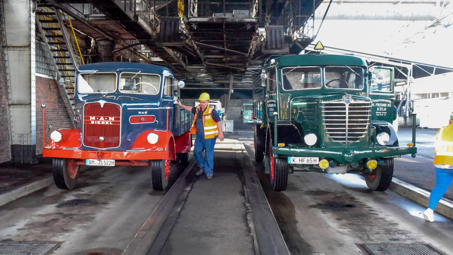 Zwei LKW aus den 1950er-Jahren stehen in der Brikettfabrik in Frechen und warten, dass sie mit Kohle beladen werden. Ein Arbeiter steht mit Helm und Warnweste neben dem linken LKW.
