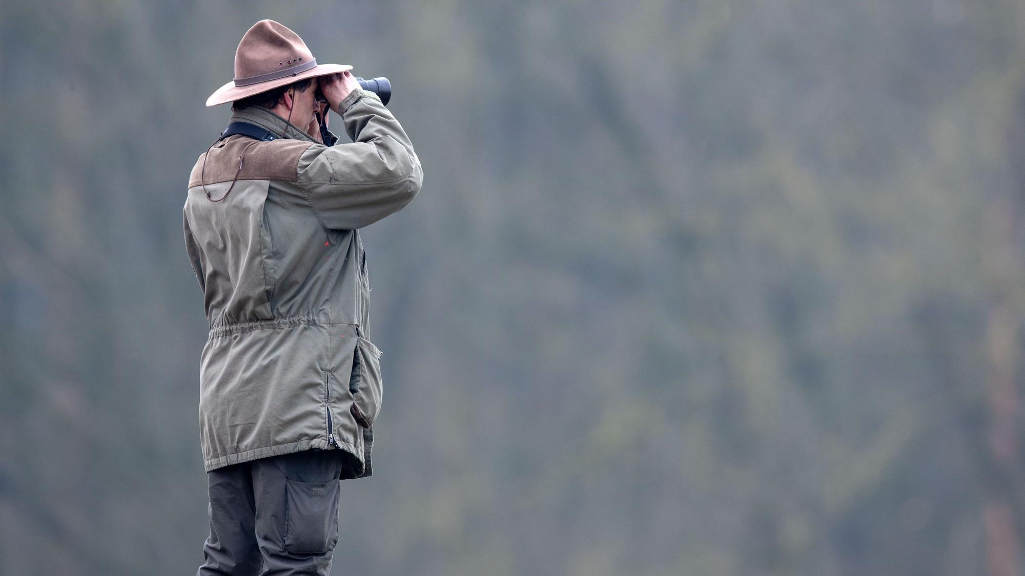 Ein Ranger schaut durch ein Fernglas auf eine tiefer liegende weite Waldfläche.
