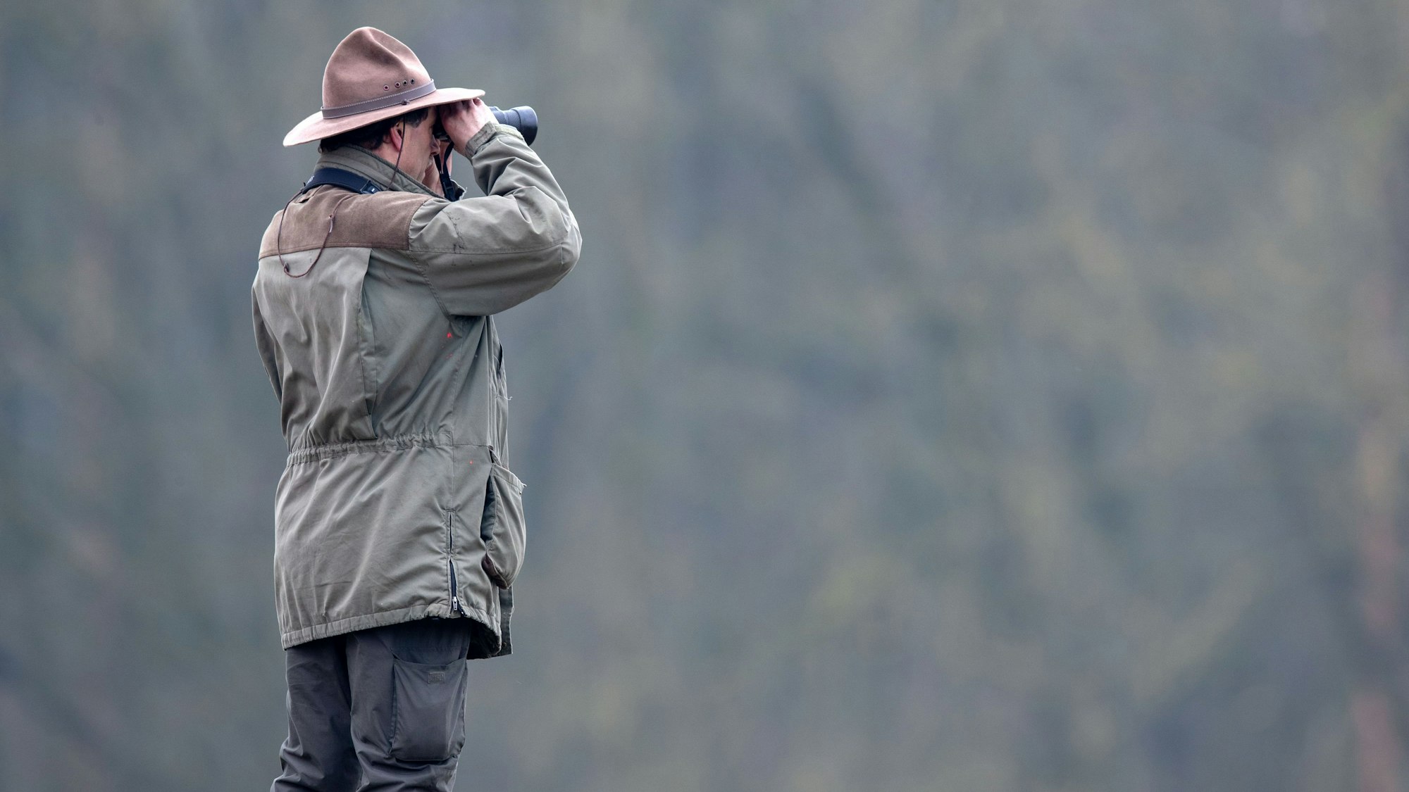 Ein Ranger schaut durch ein Fernglas auf eine tiefer liegende weite Waldfläche.