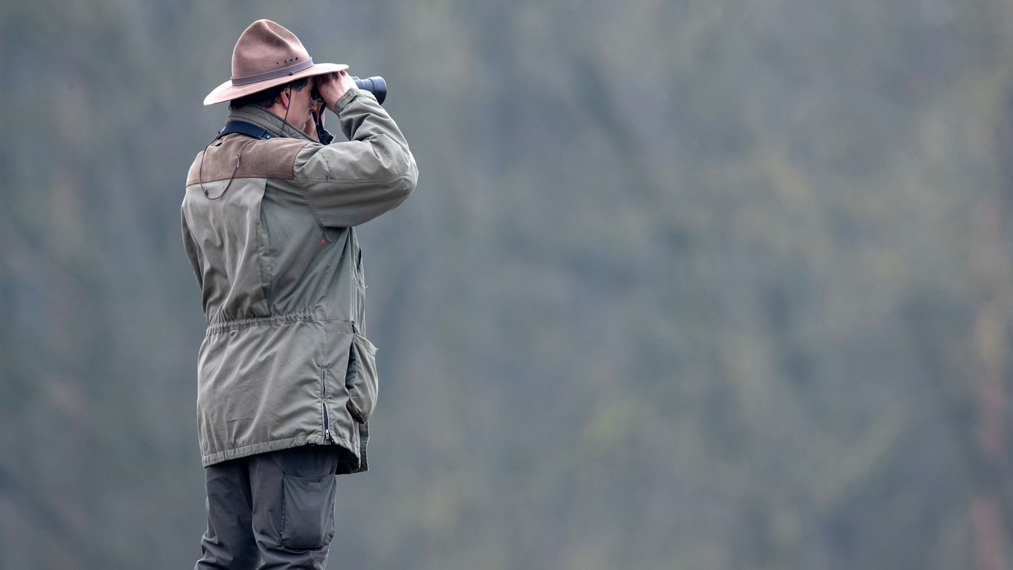 Ein Ranger schaut durch ein Fernglas auf eine tiefer liegende weite Waldfläche.