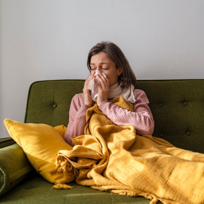 Eine Frau liegt mit einer Decke und einem Schal auf einem Sofa und putzt sich mit einem Taschentuch die Nase.