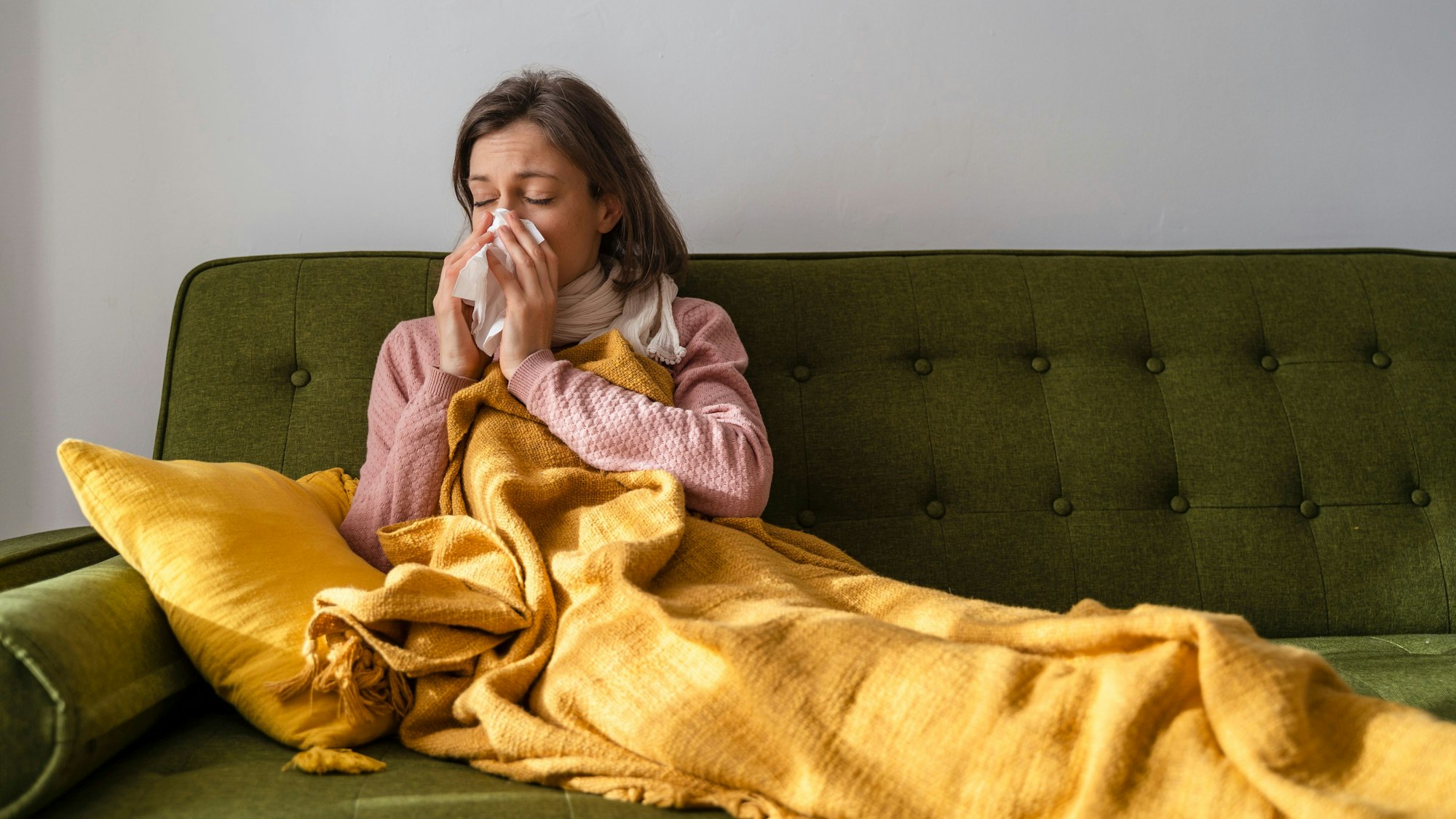 Eine Frau liegt mit einer Decke und einem Schal auf einem Sofa und putzt sich mit einem Taschentuch die Nase.