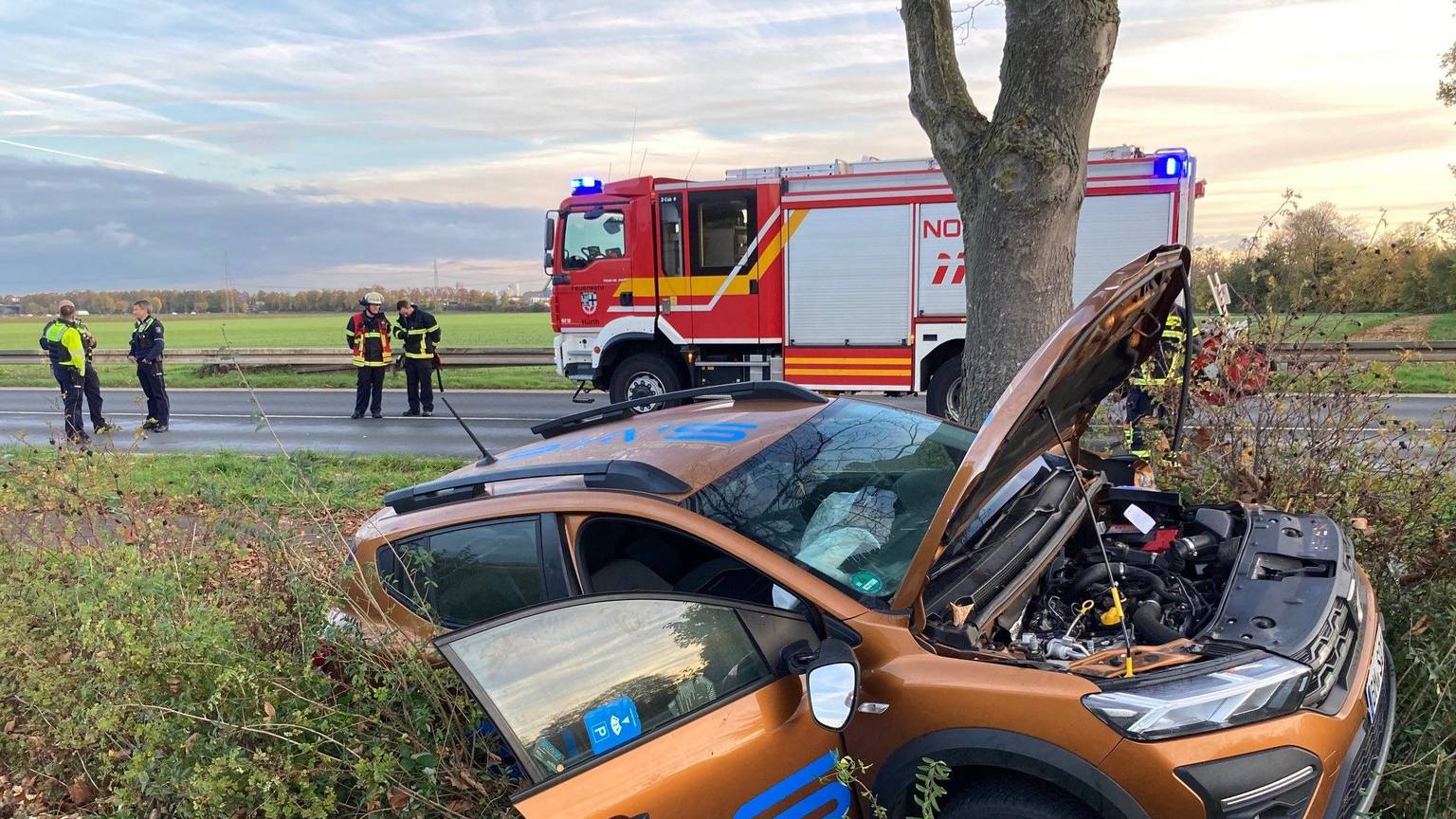 Ein Auto liegt im Straßengraben. Die Motorhaube und die Beifahrertür sind geöffnet. Auf der Straße im Hintergrund steht ein Feuerwehrauto, Polizisten und Feuerwehrleute.