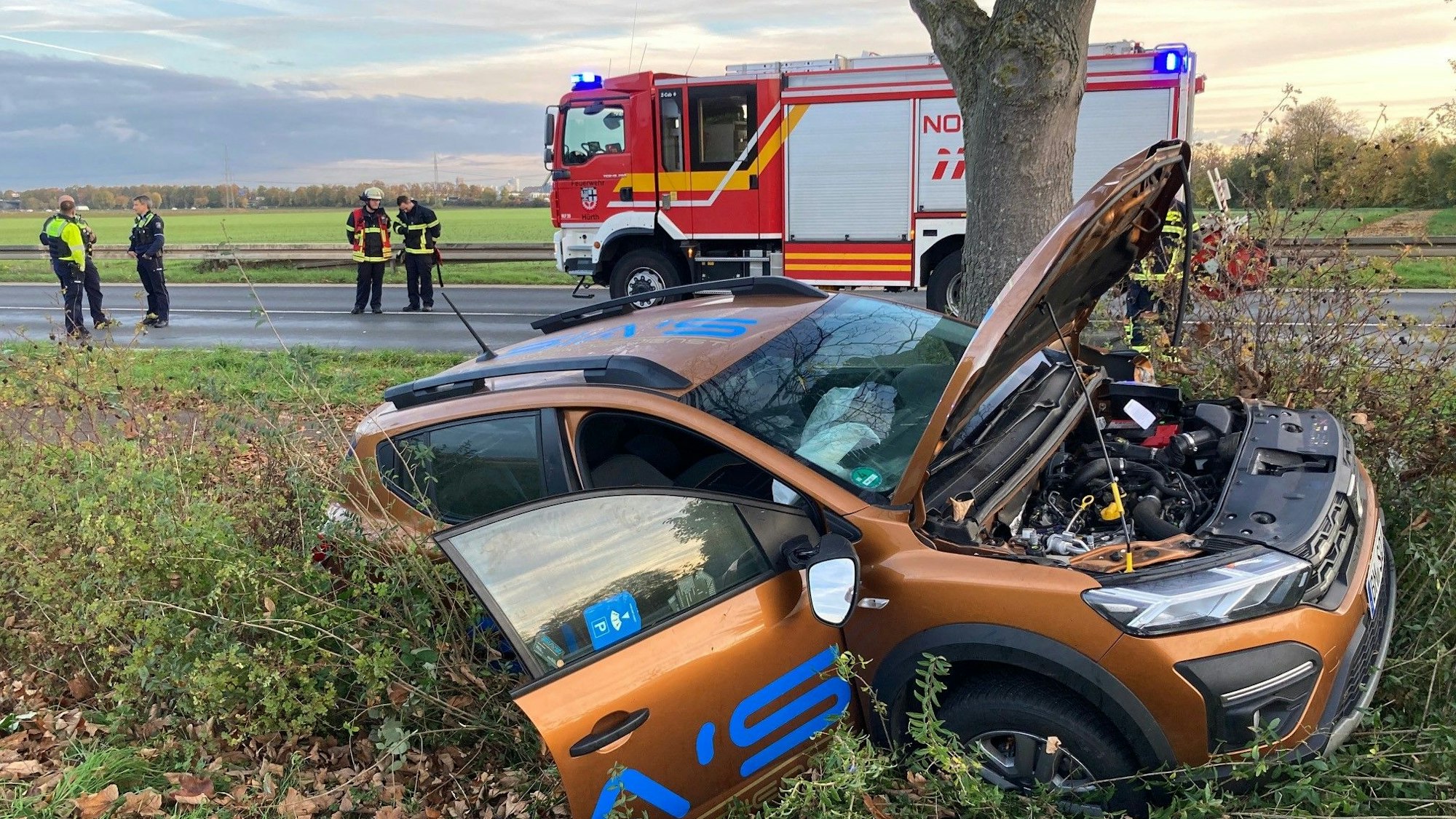 Ein Auto liegt im Straßengraben. Die Motorhaube und die Beifahrertür sind geöffnet. Auf der Straße im Hintergrund steht ein Feuerwehrauto, Polizisten und Feuerwehrleute.