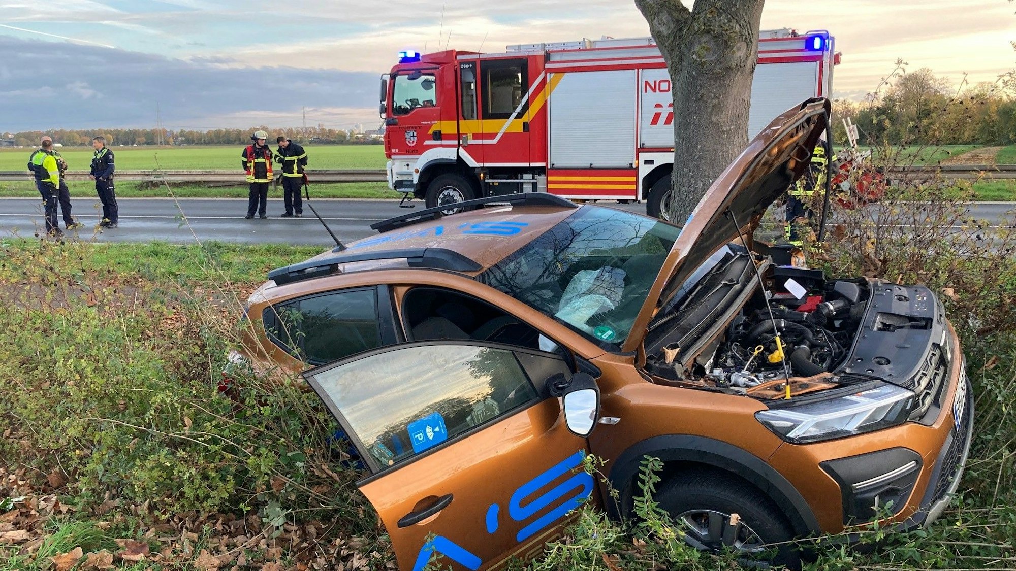 Ein Auto liegt im Straßengraben. Die Motorhaube und die Beifahrertür sind geöffnet. Auf der Straße im Hintergrund steht ein Feuerwehrauto, Polizisten und Feuerwehrleute.