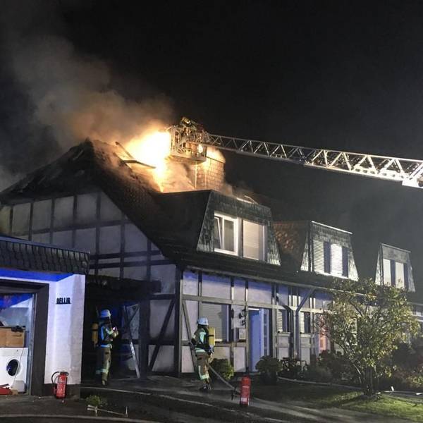 An einem Fachwerkhaus steht der Dachstuhl in Flammen. Einer von der Feuerwehr versucht aus einem Korb, der an der Drehleiter ist, das Feuer zu löschen. Zwei weiter Feuerwehrleute stehen vor einem Fachwerkhaus.