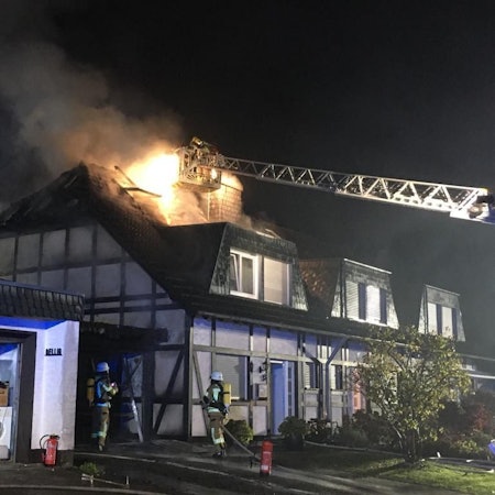 An einem Fachwerkhaus steht der Dachstuhl in Flammen. Einer von der Feuerwehr versucht aus einem Korb, der an der Drehleiter ist, das Feuer zu löschen. Zwei weiter Feuerwehrleute stehen vor einem Fachwerkhaus.