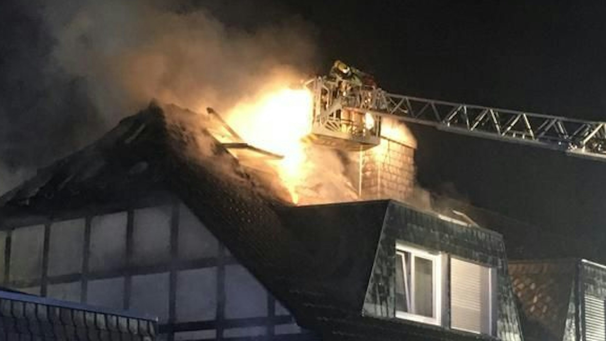 An einem Fachwerkhaus steht der Dachstuhl in Flammen. Einer von der Feuerwehr versucht aus einem Korb, der an der Drehleiter ist, das Feuer zu löschen. Zwei weiter Feuerwehrleute stehen vor einem Fachwerkhaus.