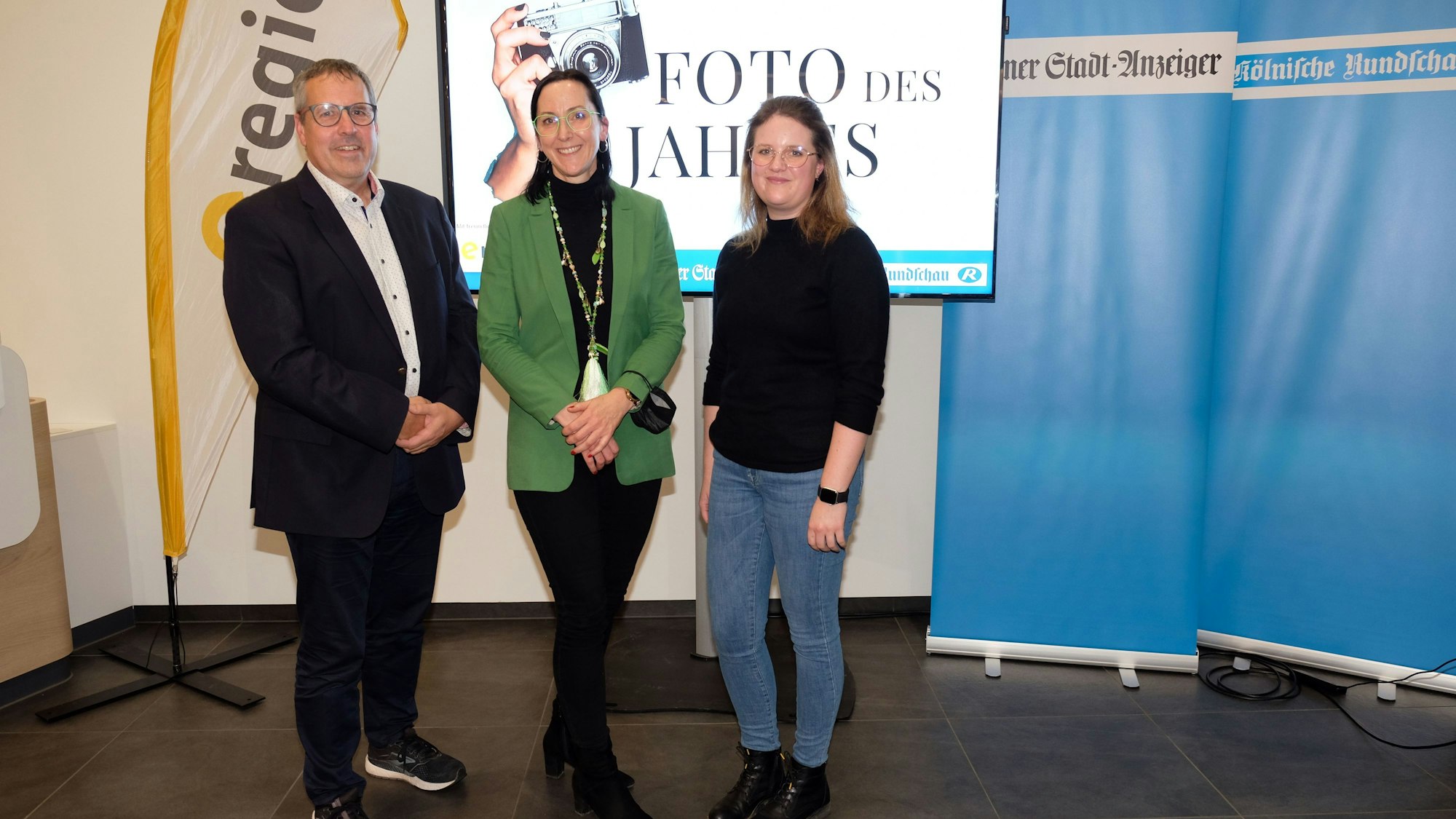 Ein Mann und zwei Frauen stehen vor einem Bildschirm mit der Aufschrift „Foto des Jahres“. Daneben stehen Banner vom „Kölner Stadt-Anzeiger“ und „Kölnische Rundschau“.