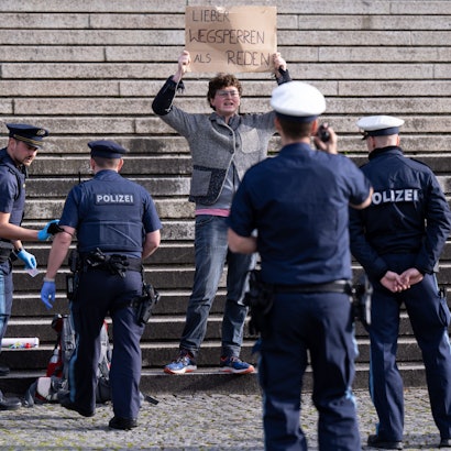 Ein Klimaaktivist steht vor der bayerischen Staatskanzlei und halten ein Schild mit der Aufschrift„ Lieber wegsperren als reden“  in den Händen. Um ihn herum stehen Polizisten.