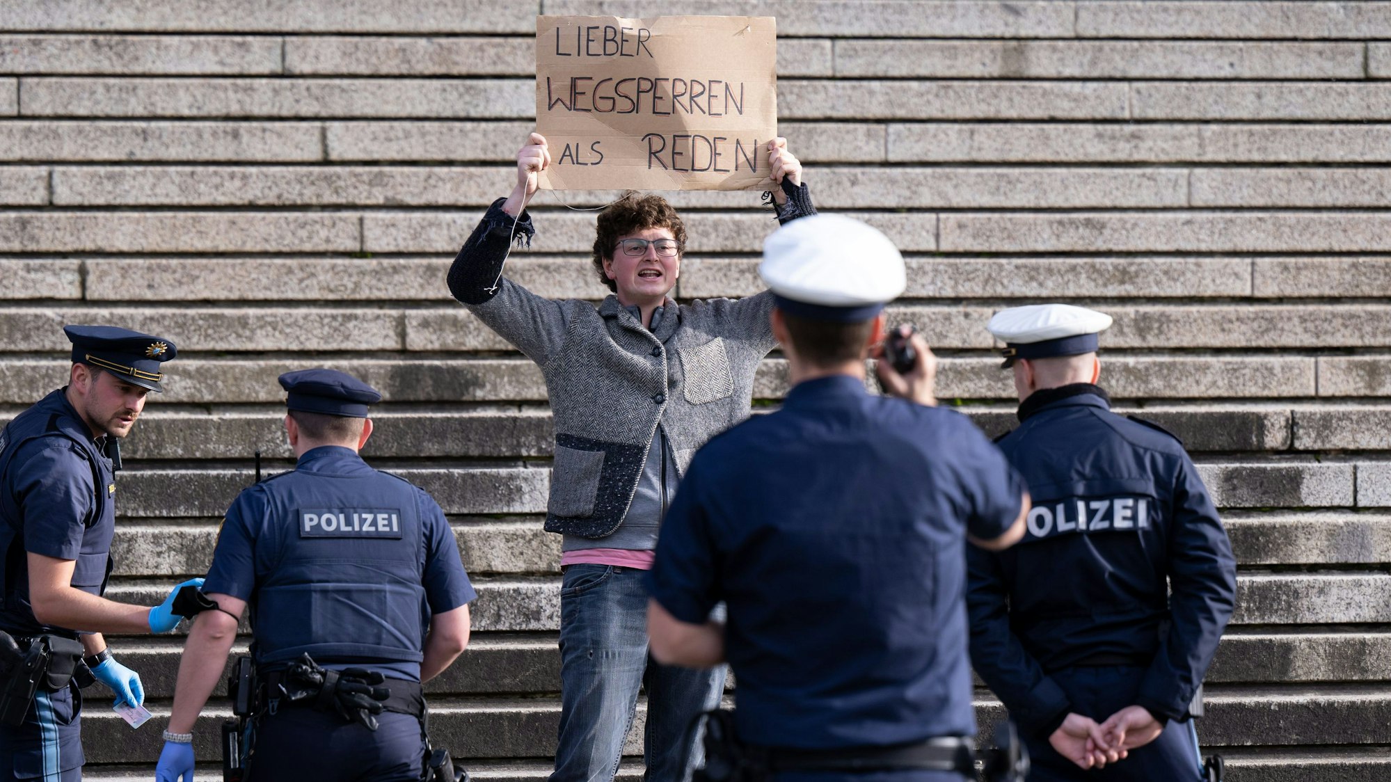 Ein Klimaaktivist steht vor der bayerischen Staatskanzlei und halten ein Schild mit der Aufschrift„ Lieber wegsperren als reden“ in den Händen. Um ihn herum stehen Polizisten.