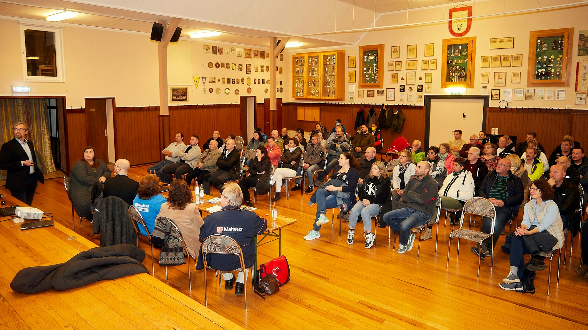 Rund 55 Personen sitzen in mehreren Reihen auf Stühlen. Davor sitzen fünf Männer und Frauen an einem Tisch.