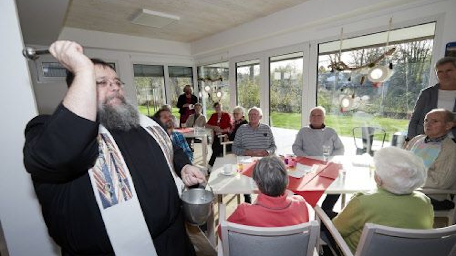Pater Georg segnet einen Raum mit Weihwasser. Im Hintergrund sitzen Seniorinnen und Senioren an Tischen vor einer Fensterfront.