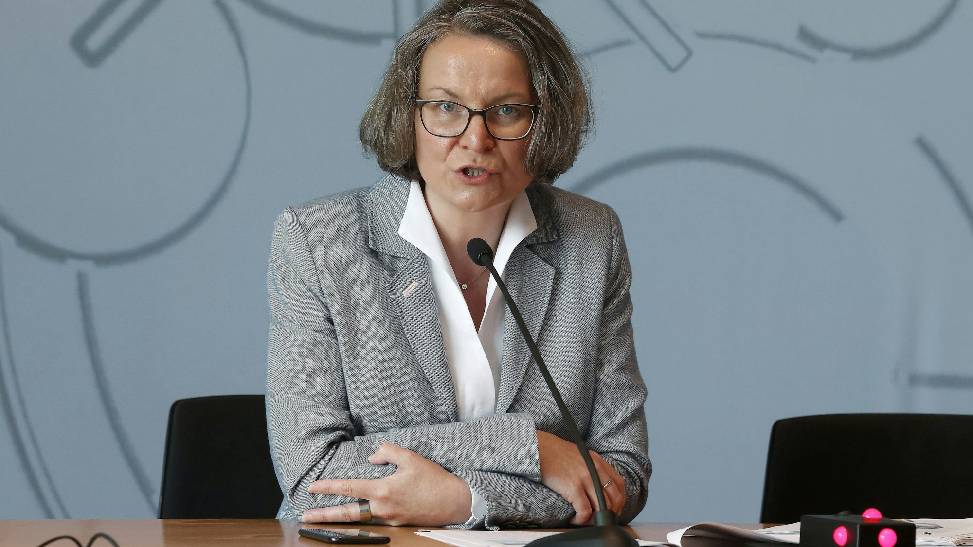 NRW-Ministerin Ina Scharrenbach (CDU) bei einer Pressekonferenz. Sie sitzt an einem Tisch, vor ihr ein schmales Mikrofon und hinter ihr eine graue Wand.