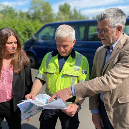 Eine Frau und zwei Männer stehen auf einem Parkplatz und schauen auf eine Karte.