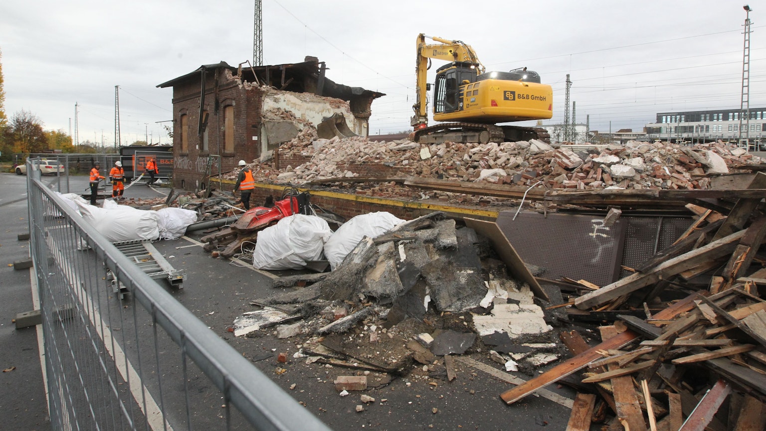 Ein gelber Bagger steht auf den Trümmern der Lagerhalle. Im Hintergrund ist noch ein Teil des Gebäudes aus Backsteinen zu sehen. Männer in orangen Sicherheitswesten arbeiten auf der Baustelle.
