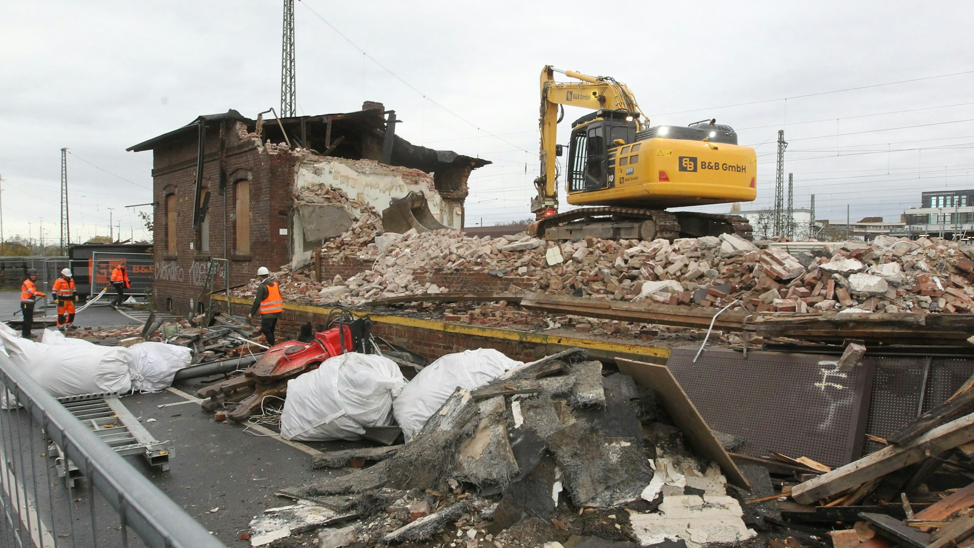 Ein gelber Bagger steht auf den Trümmern der Lagerhalle. Im Hintergrund ist noch ein Teil des Gebäudes aus Backsteinen zu sehen. Männer in orangen Sicherheitswesten arbeiten auf der Baustelle.