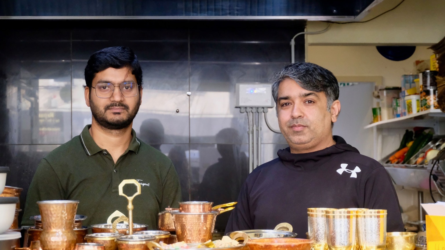 Kabil Kataria (links) steht mit einem Mitarbeitern hinter der Theke in seinem neuen Imbiss Curry Up. Vor ihm stehen verschiedene Becher und Schalen in silberner oder Aluminium-Optik. Rechts hinter ihnen stehen viele verschiedene Gewürze in einem Regal.