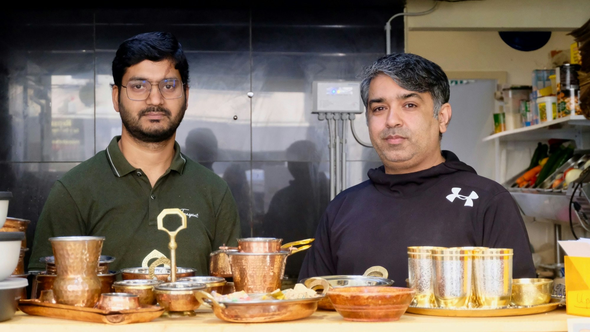 Kabil Kataria (links) steht mit einem Mitarbeitern hinter der Theke in seinem neuen Imbiss Curry Up. Vor ihm stehen verschiedene Becher und Schalen in silberner oder Aluminium-Optik. Rechts hinter ihnen stehen viele verschiedene Gewürze in einem Regal.