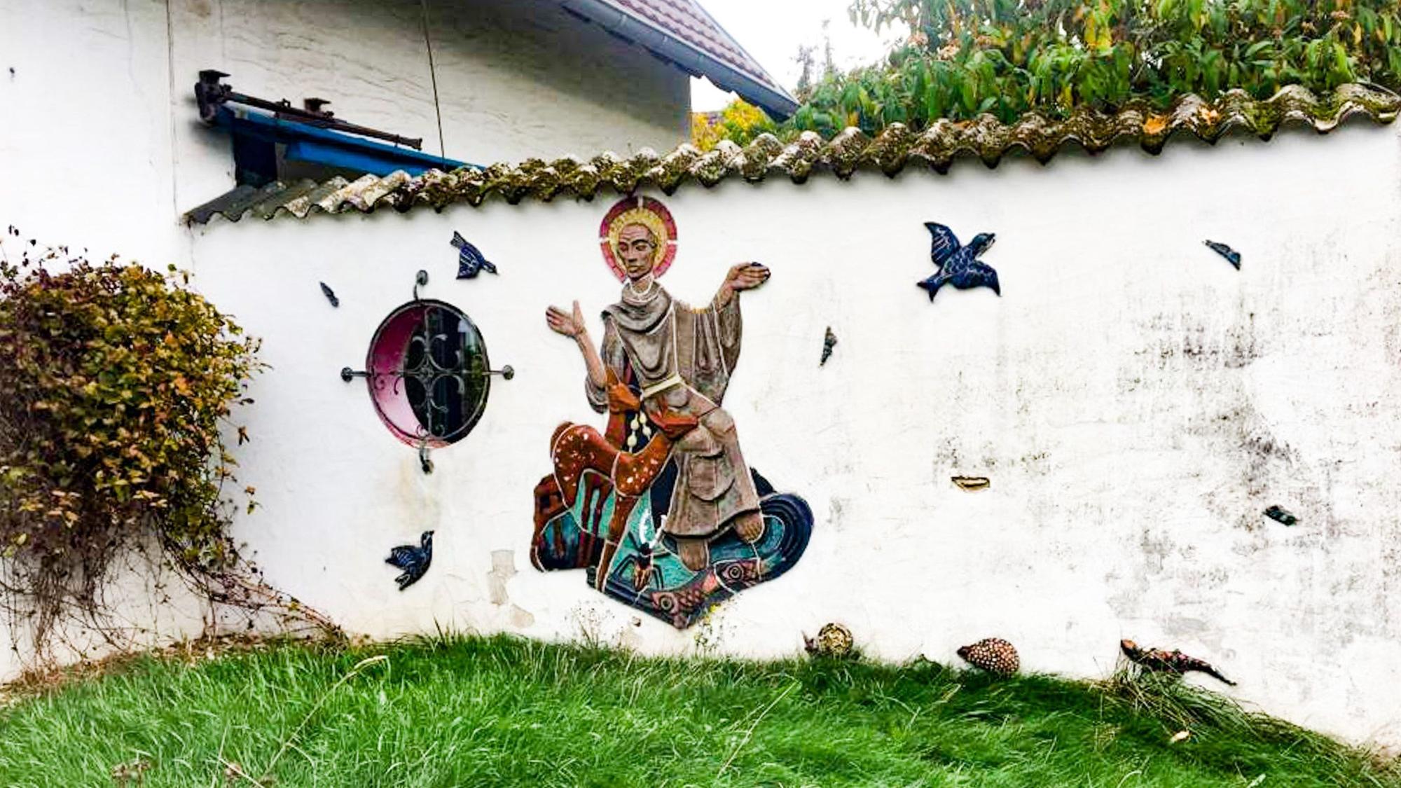 An einer Gartenmauer ist aus Keramik der Heilige Franz von Assisi dargestellt mit Tauben, Igel und anderen Tieren.