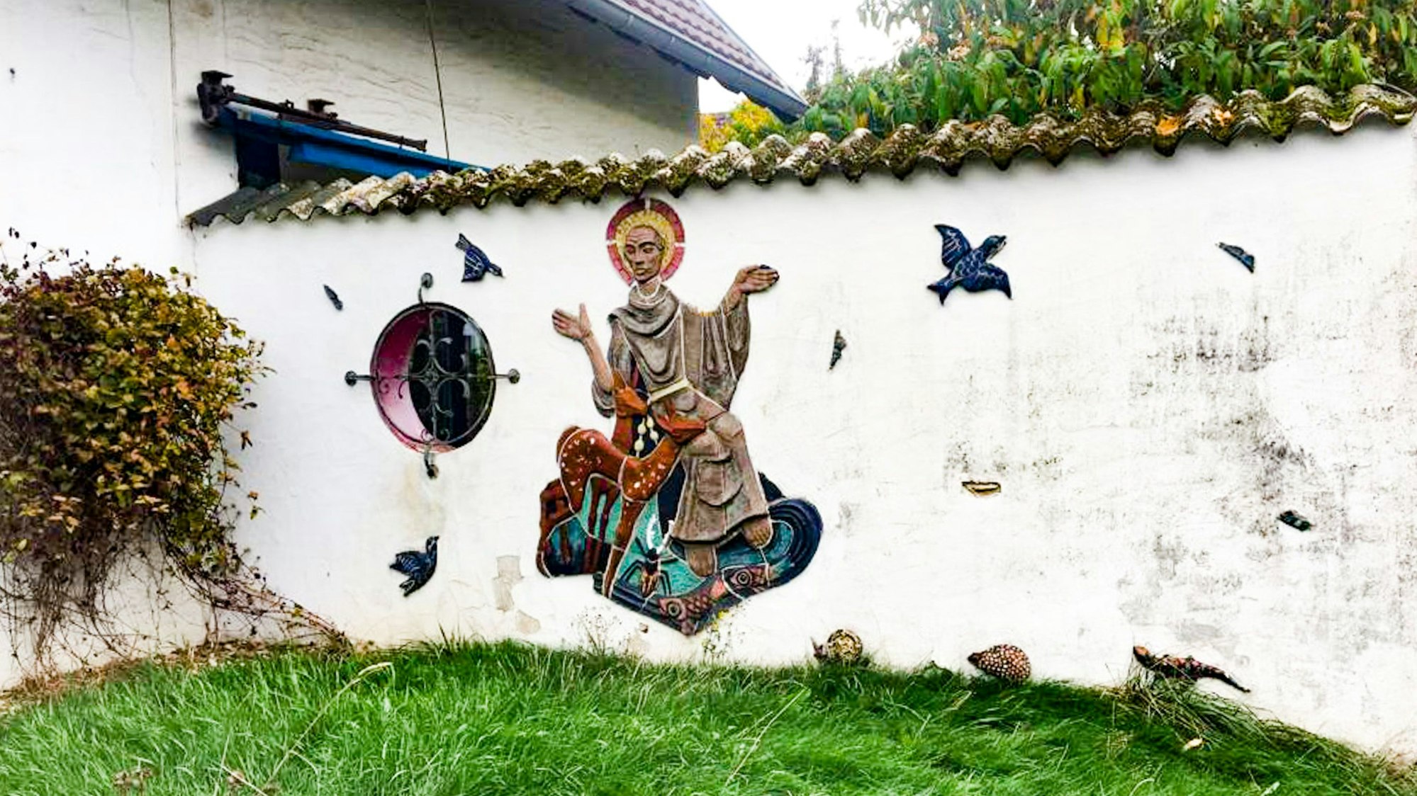 An einer Gartenmauer ist aus Keramik der Heilige Franz von Assisi dargestellt mit Tauben, Igel und anderen Tieren.