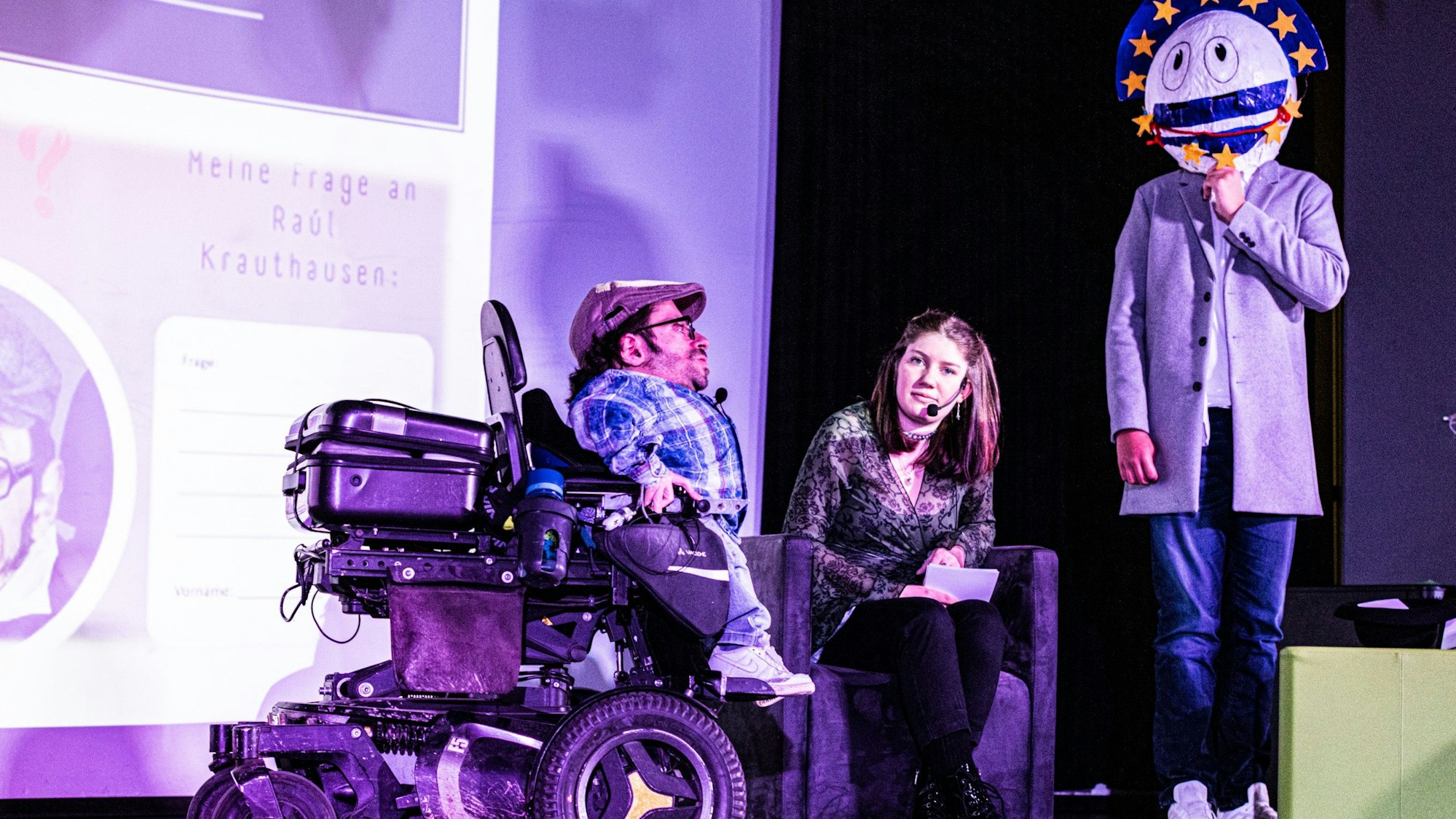 Preisverleihung auf der Bühne in Bornheim: Raúl Aguayo-Krauthausen in seinem Rollstuhl mit der Moderatorin sowie dem Schulmaskottchen "Borni".