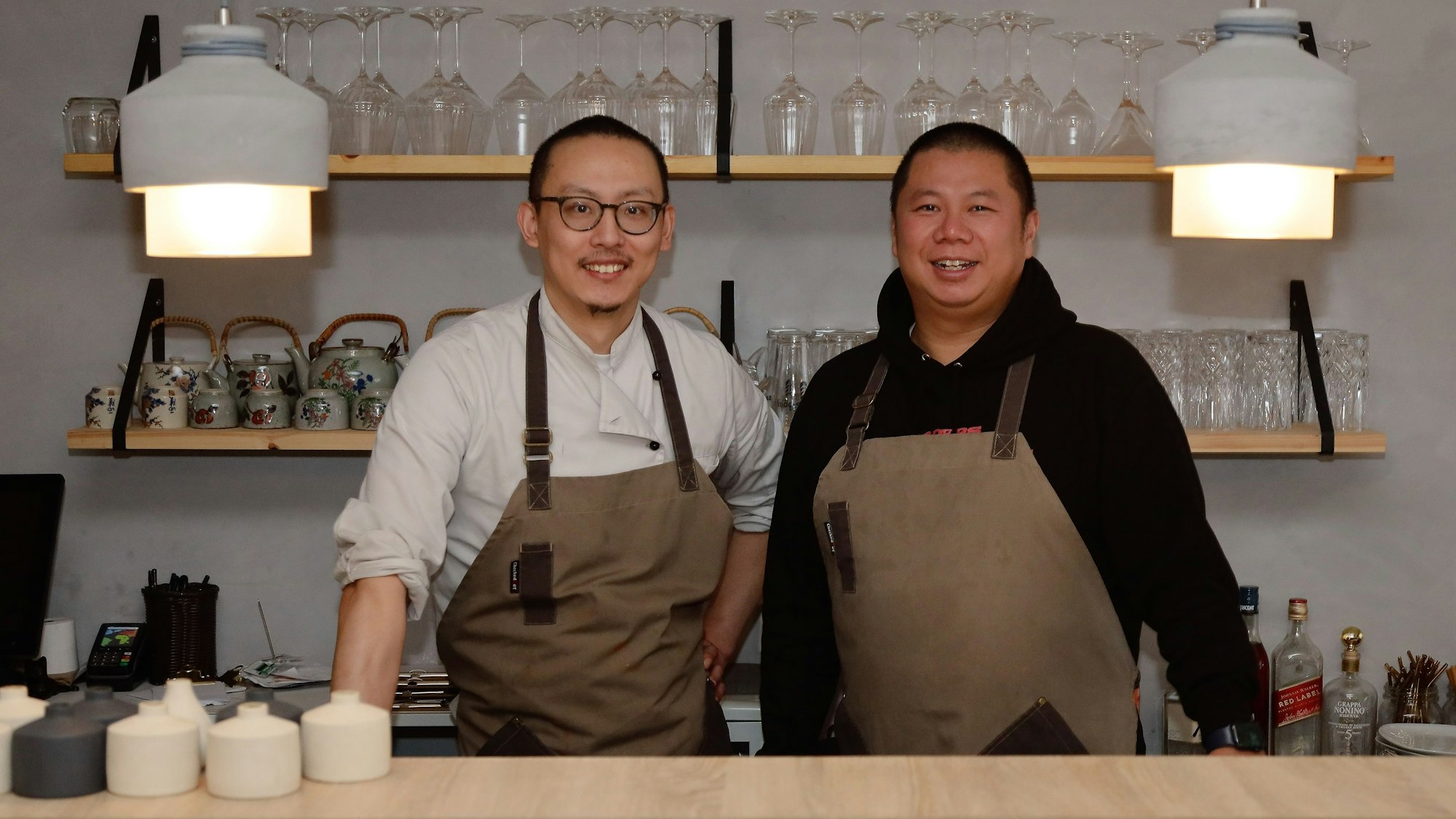 Koch Juju und der neue Serviceleiter Tutu stehen hinter einem Tresen.