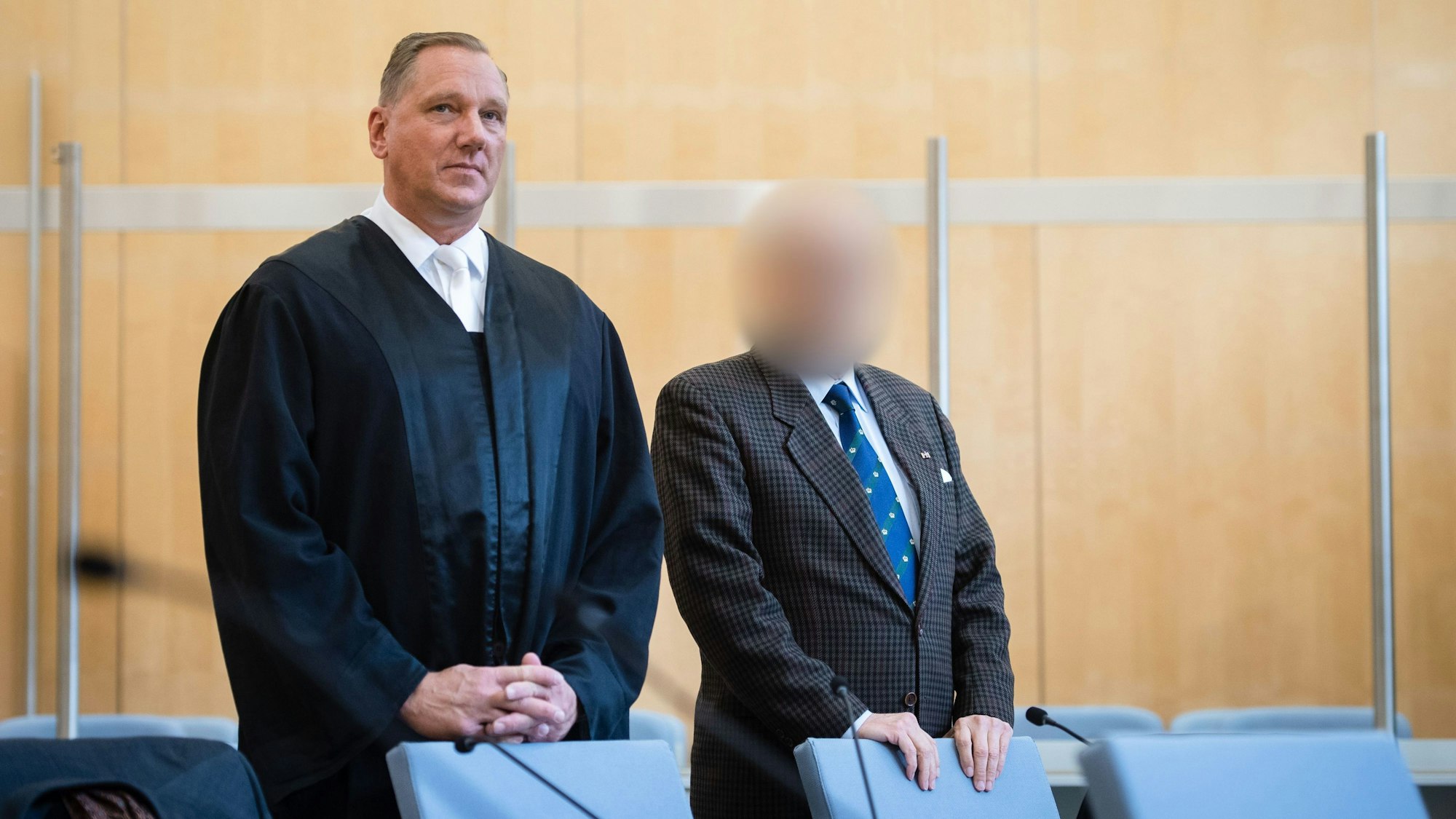 Blick in den Düsseldorfer Gerichtssaal, ein Angeklagter steht im Anzug hinter einem Stuhl, auf dem er lehnt, sein Gesicht ist nicht zu erkennen. Sein Verteidiger steht neben ihm.